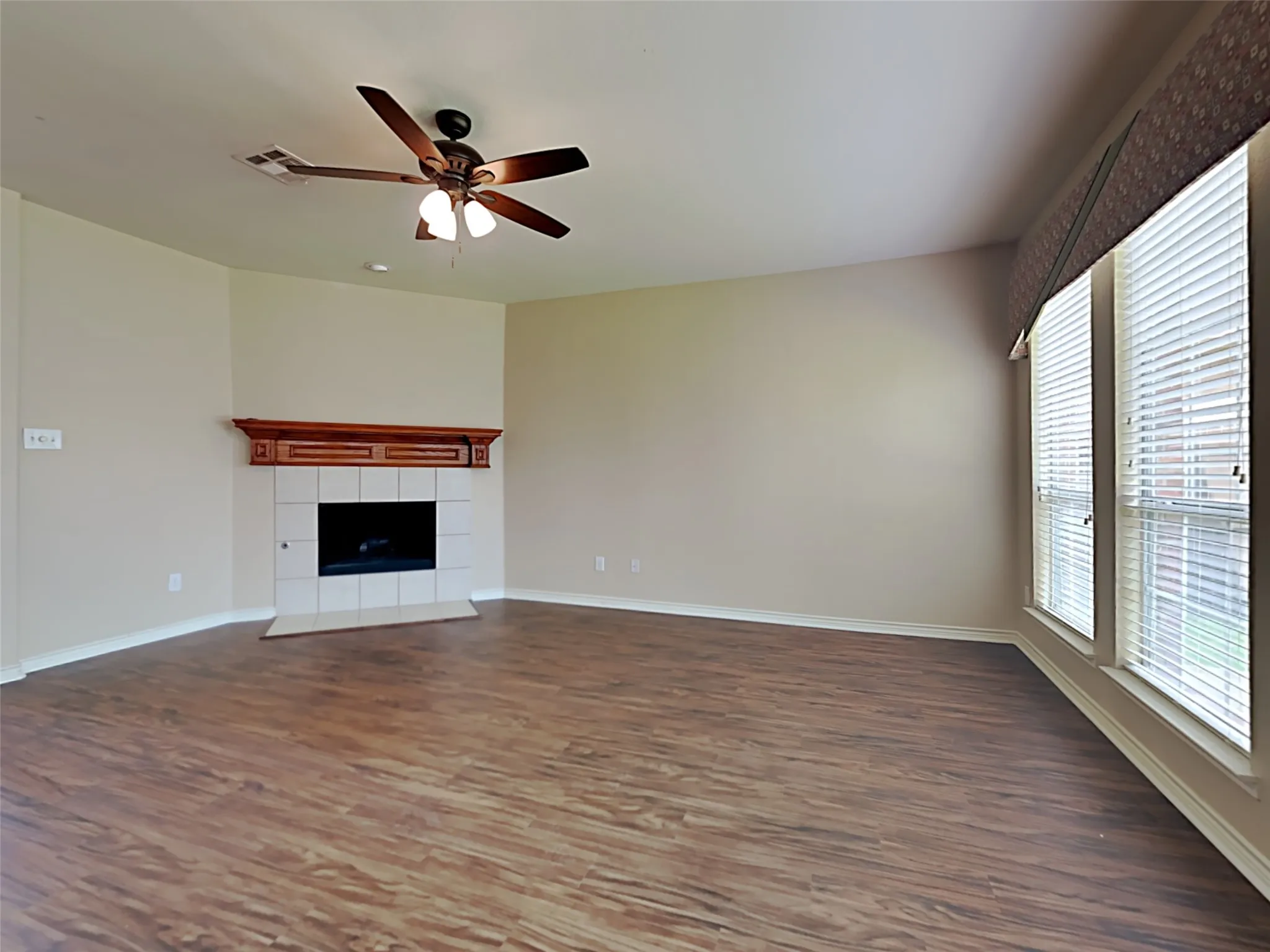Spacious living area featuring wood-finish flooring, a corner fireplace with tile surround and wood mantel, and multiple windows with blinds