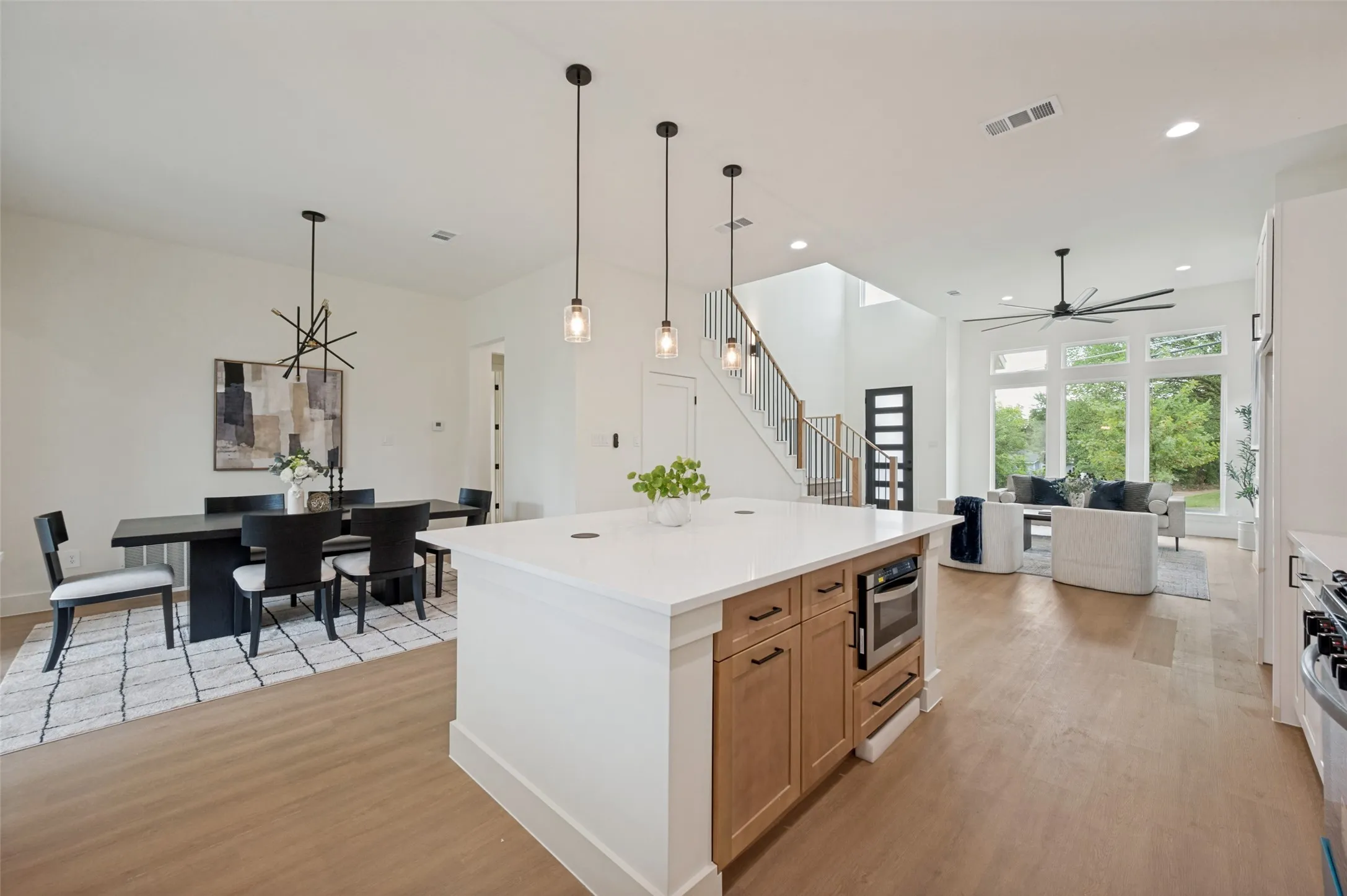 Expansive open-concept living area featuring light wood-finish flooring, a kitchen island with stone countertop, built-in microwave, and pendant lighting, and a living room with tall windows