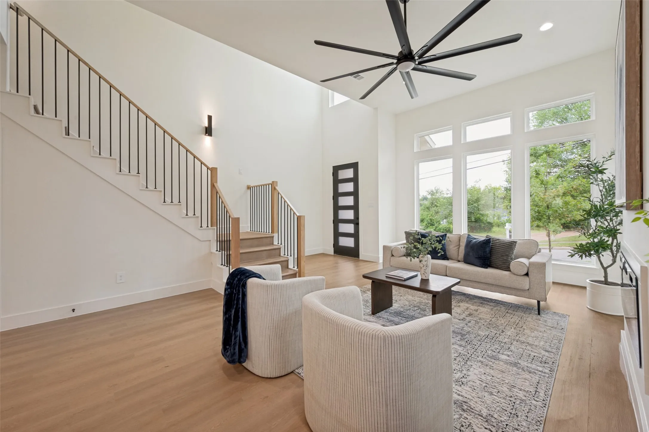 Two-story living area with wood-finish flooring, a contemporary staircase featuring wood handrails and black balusters, a dark ceiling fan, and large windows providing ample natural light