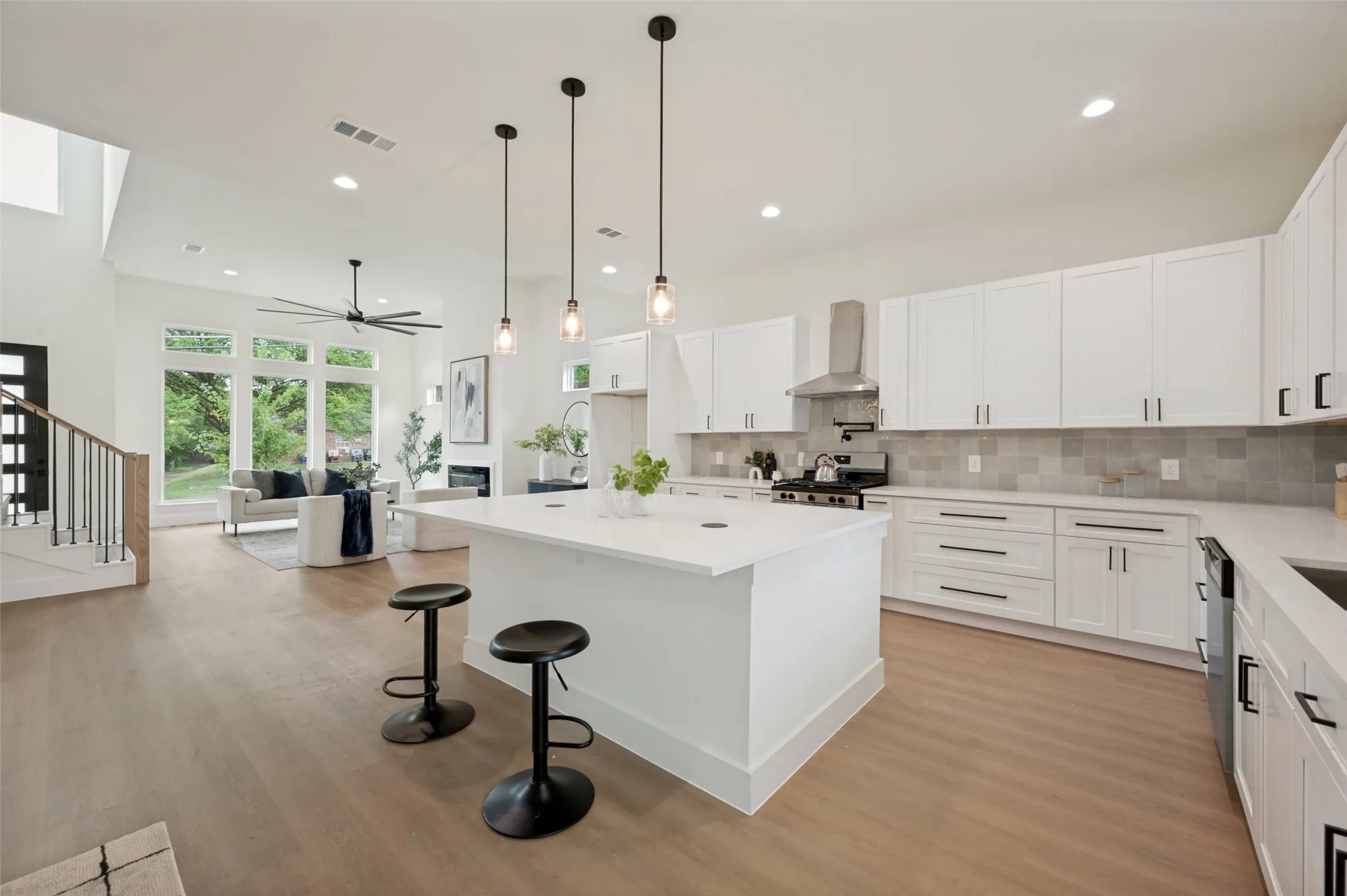 Open-concept design featuring wood-finish flooring, a kitchen island with seating, white shaker cabinetry, a stainless steel range hood, and large windows