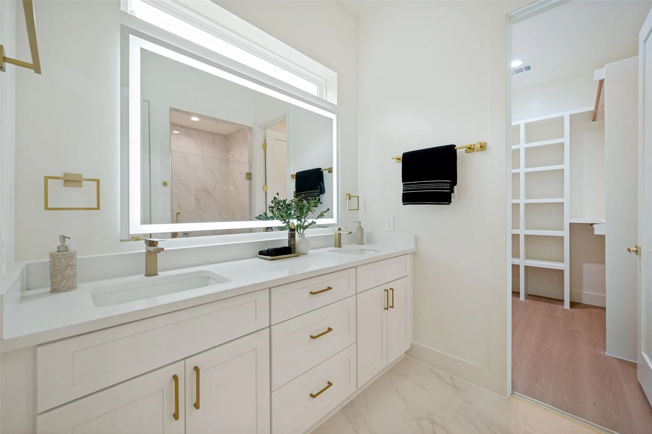 Dual vanity with white cabinetry, gold-finish hardware, and an illuminated mirror
