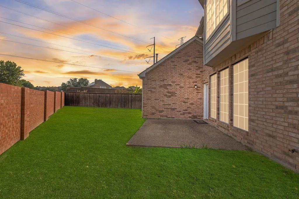 Expansive backyard featuring a manicured lawn, concrete patio, brick privacy wall, and multi-pane windows