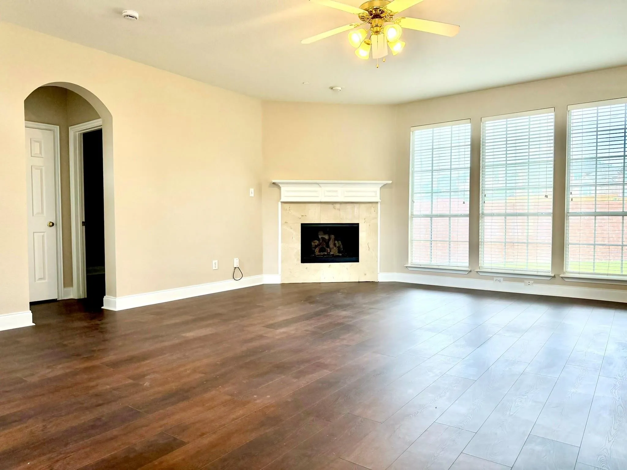 Wood-finish flooring, three large windows with blinds, an archway, a built-in fireplace with a mantel, and a ceiling fan with integrated lighting