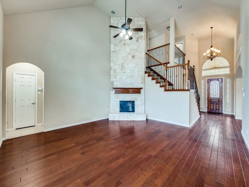 Vaulted ceiling with a ceiling fan, stone-clad fireplace with wood mantle, wood-finish flooring, two-story staircase with dark wrought iron spindles, and an arched entry door with decorative glass