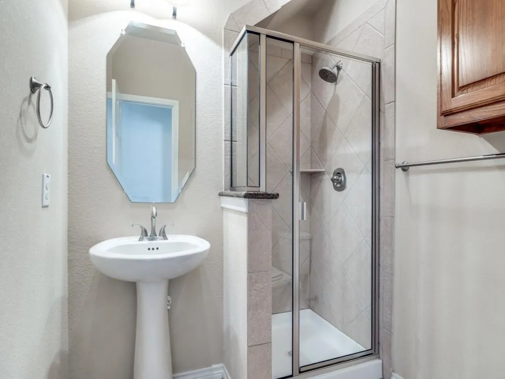 Pedestal sink with chrome fixtures, glass-enclosed shower with tiled surround, wall-mounted wood-finish cabinetry, and an octagonal framed mirror