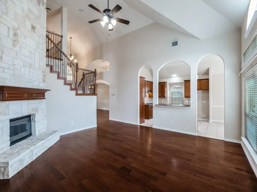 Hardwood-finish flooring, stone-clad fireplace with wood mantel, vaulted ceilings, arched doorways, and a ceiling fan with integrated lighting
