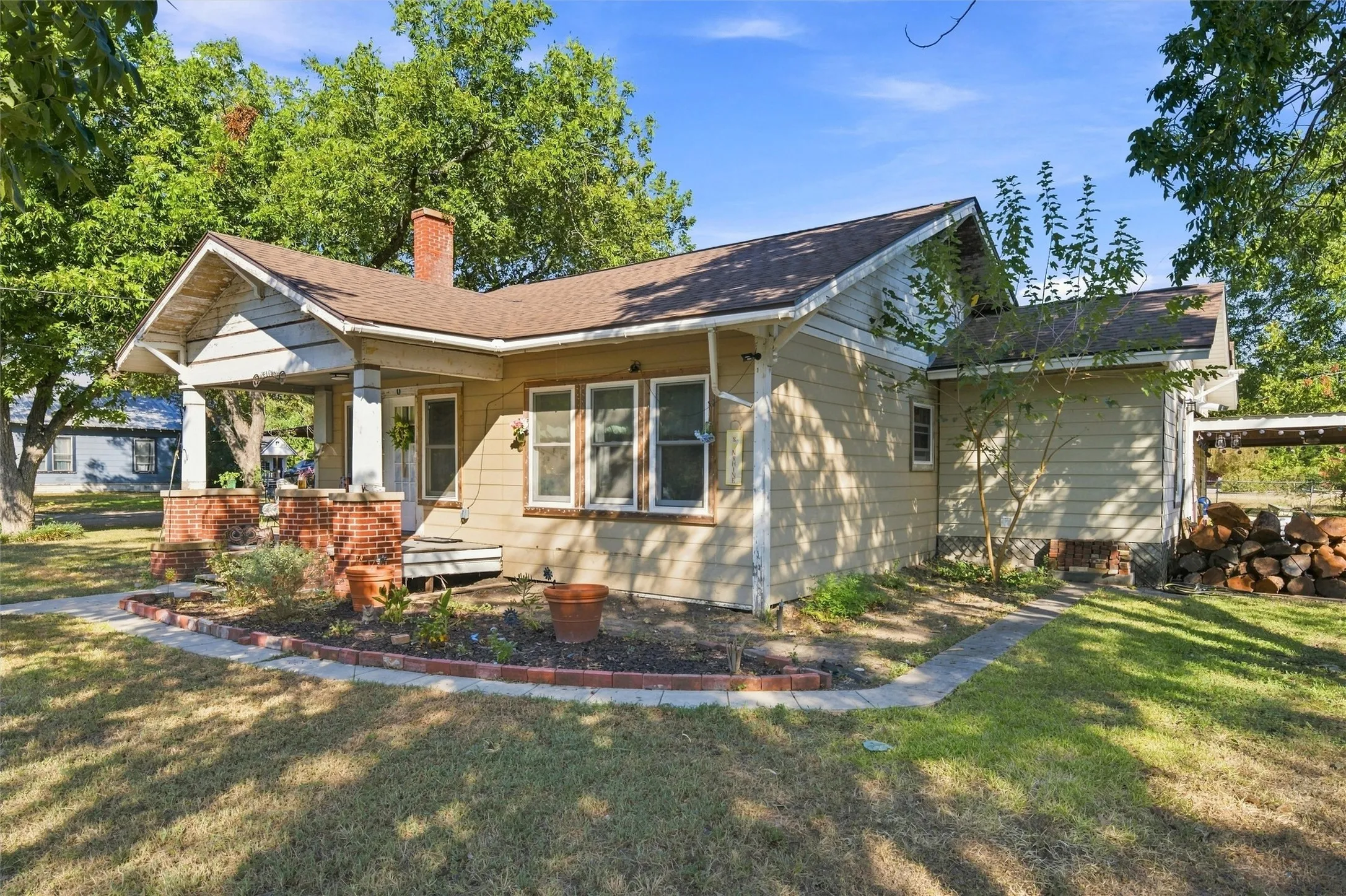 Front porch with brick piers, wood siding exterior, shingled roof, and mature trees