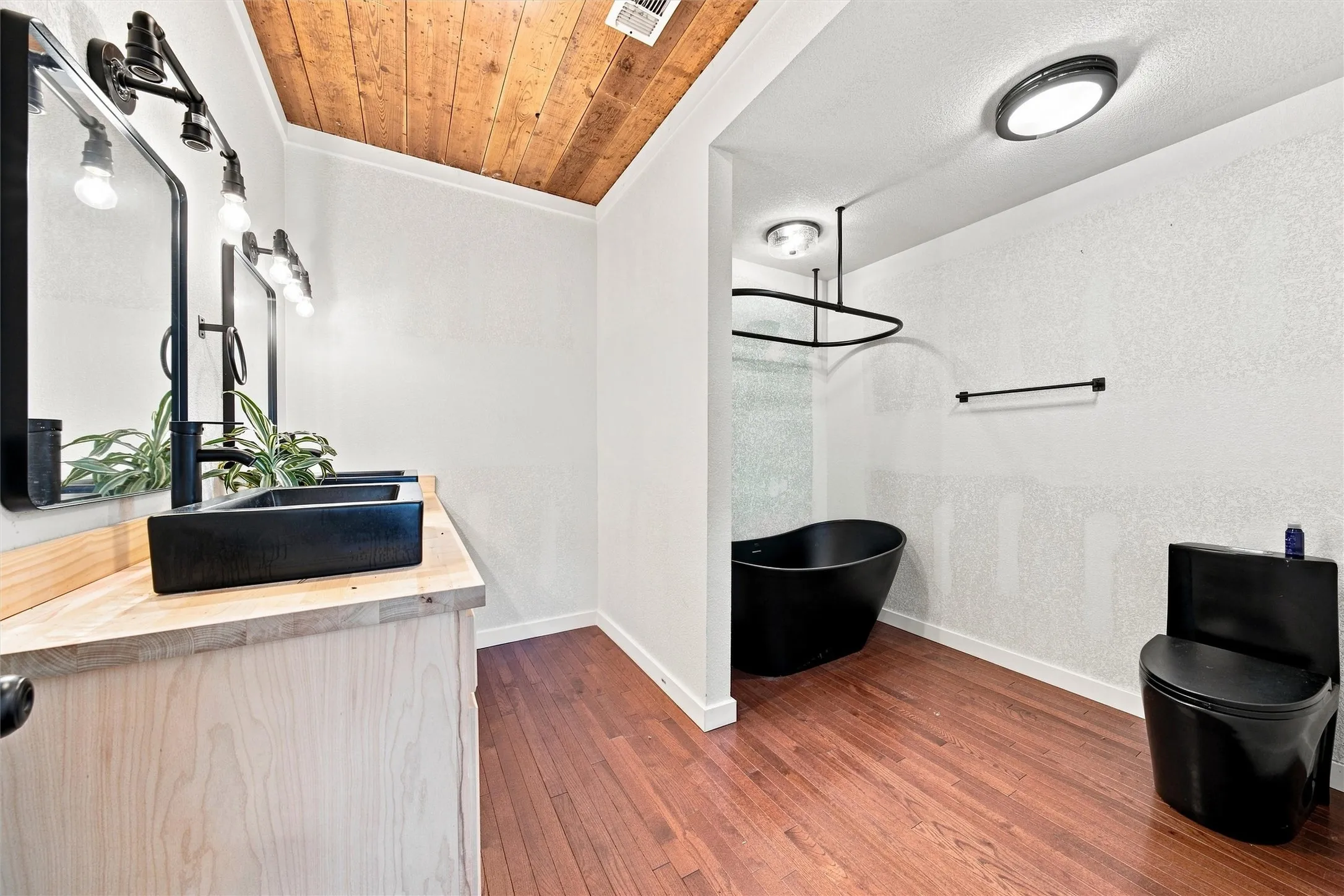 Wood-paneled ceiling, dark wood-finish flooring, light wood vanity, black vessel sink, and black freestanding bathtub