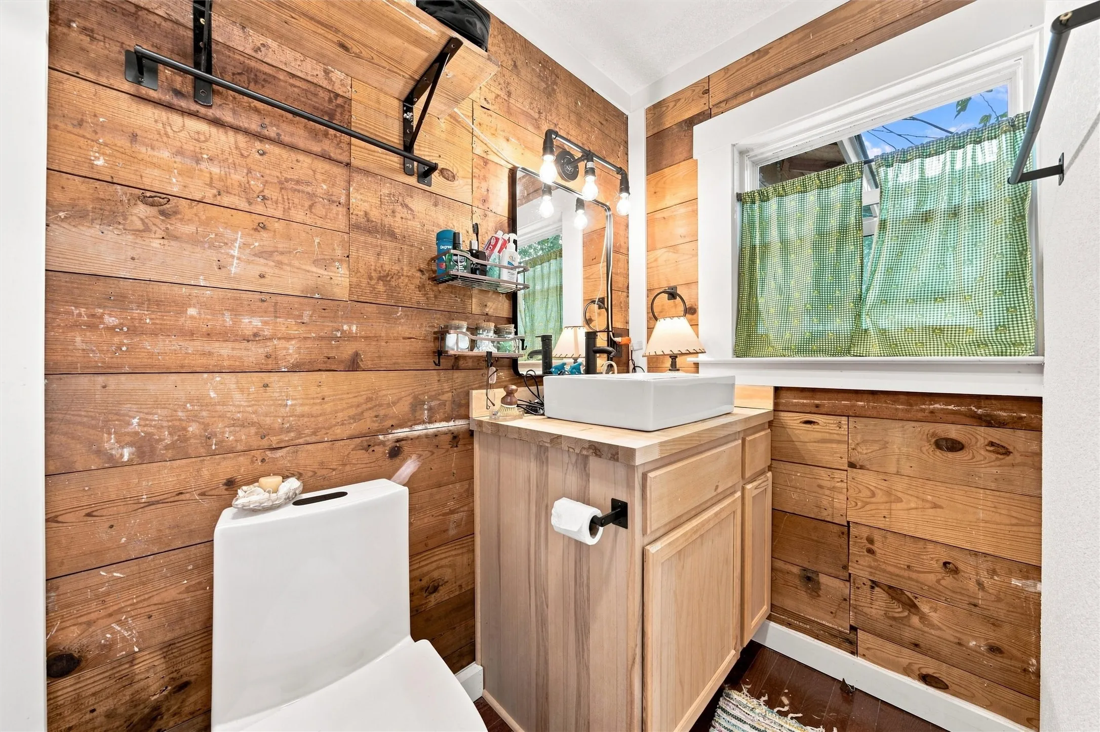 Wood-paneled wall accents, vessel sink, vanity with storage, dark wood-finish flooring, and a window with natural light