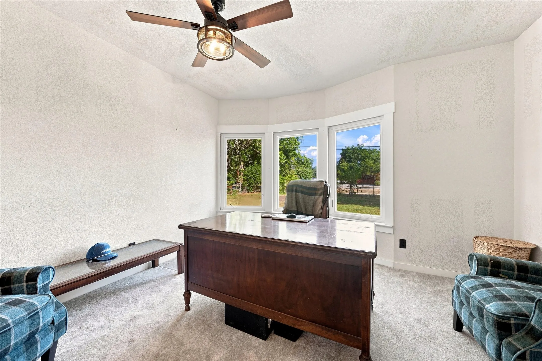 Bay window with three pane windows, ceiling fan with light fixture, light-colored carpet, and textured walls