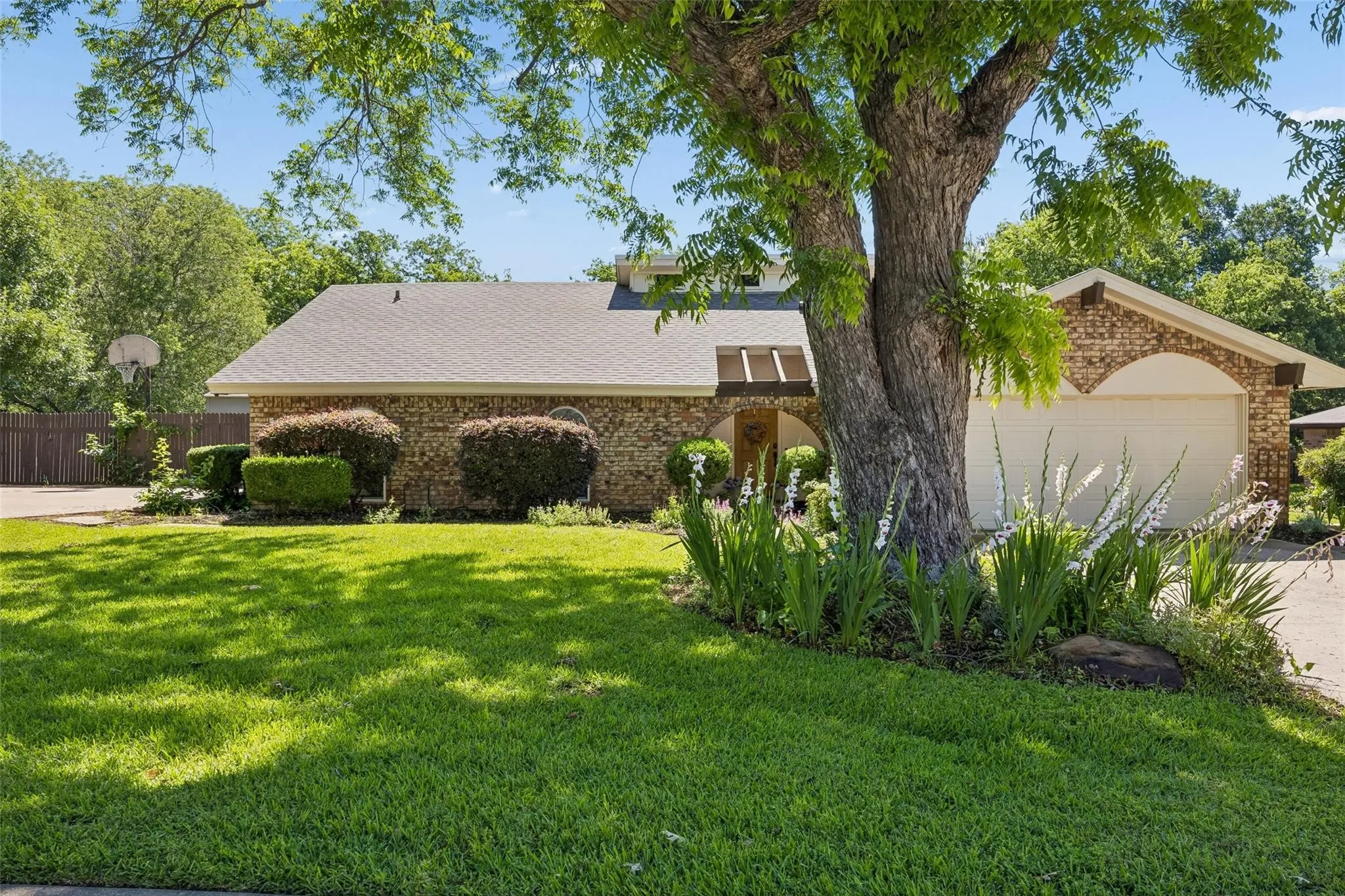 Brick exterior home featuring a two-car garage, established front landscaping, and a mature shade tree
