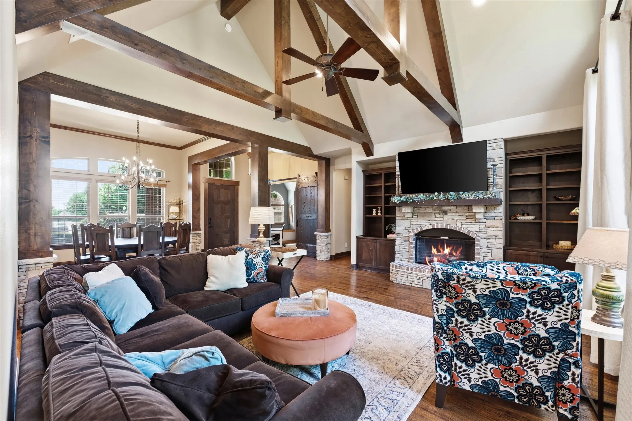 Vaulted ceiling with exposed wood beams, hand scraped hardwood floors, stone-clad gas fireplace with mantel, built-in shelving, and large picture windows