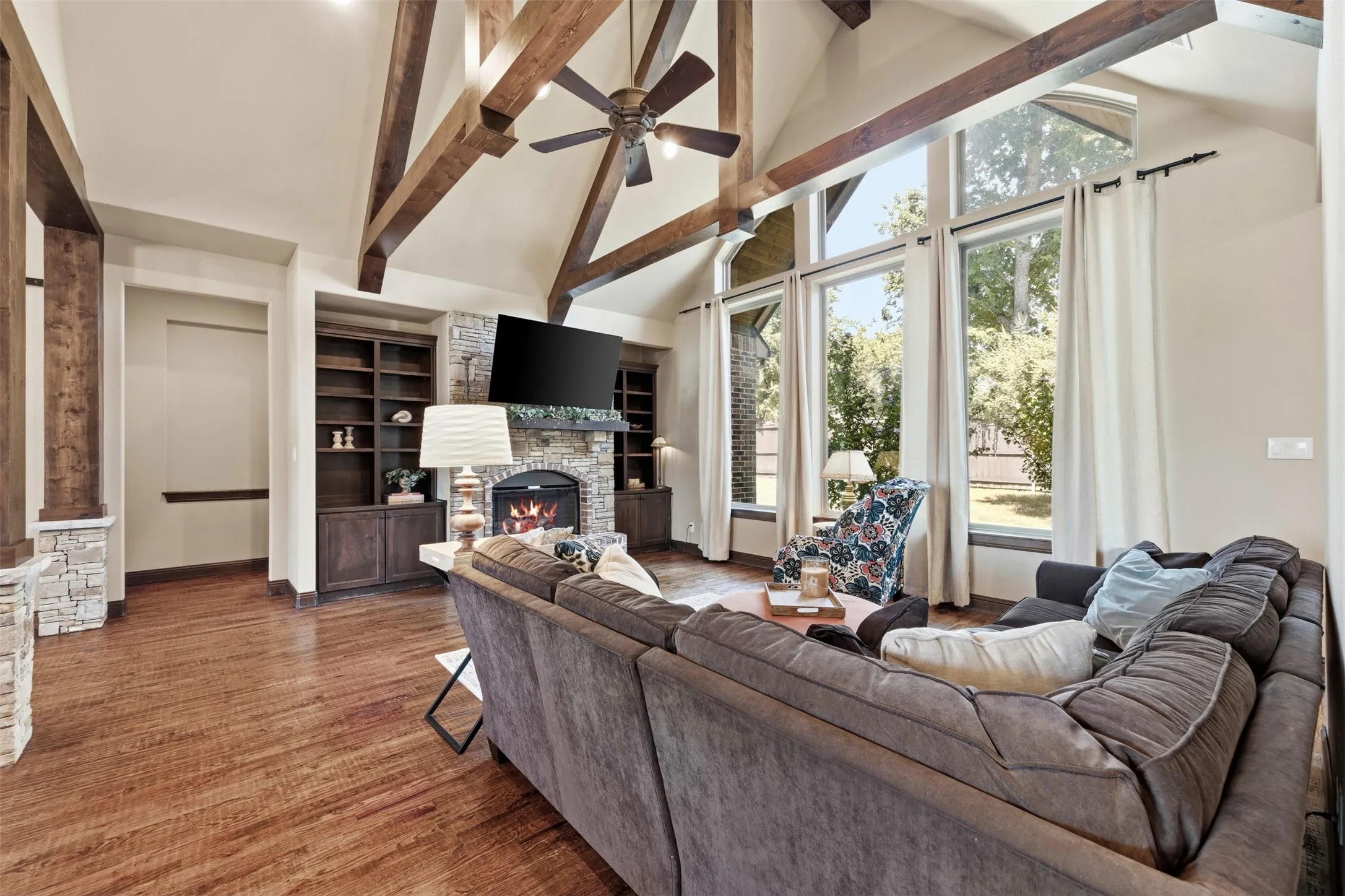 Vaulted ceiling with exposed wood beams, hand scraped hardwood floors, stone-clad gas fireplace with mantel, built-in shelving, and large picture windows