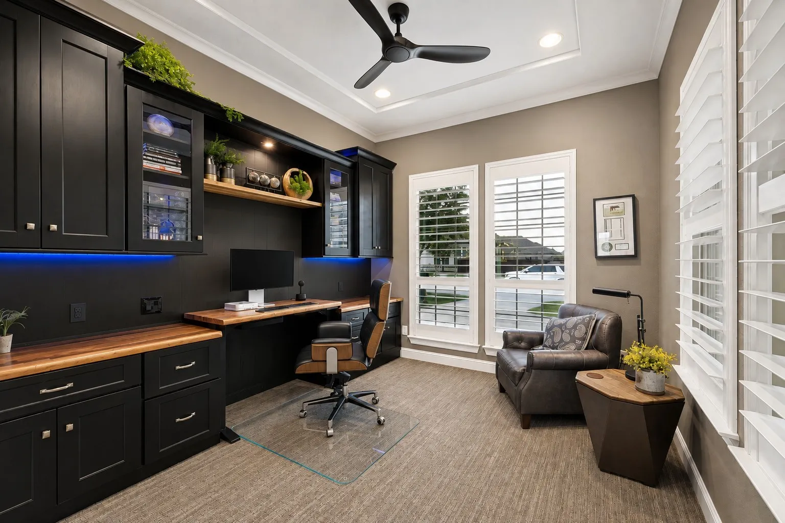 A sophisticated private office designed to make working from home feel effortless, featuring striking custom cabinetry with built-in storage, rich walnut countertops, and an adjustable sit-to-stand desk for the flexibility to work seated or standing. Decorative accent lighting, a designer ceiling fan, luxurious low-profile carpet, and plantation shutters elevate the space, while elegant glass French doors provide privacy without sacrificing natural light. A beautifully appointed workspace where luxury, comfort, and functionality come together.