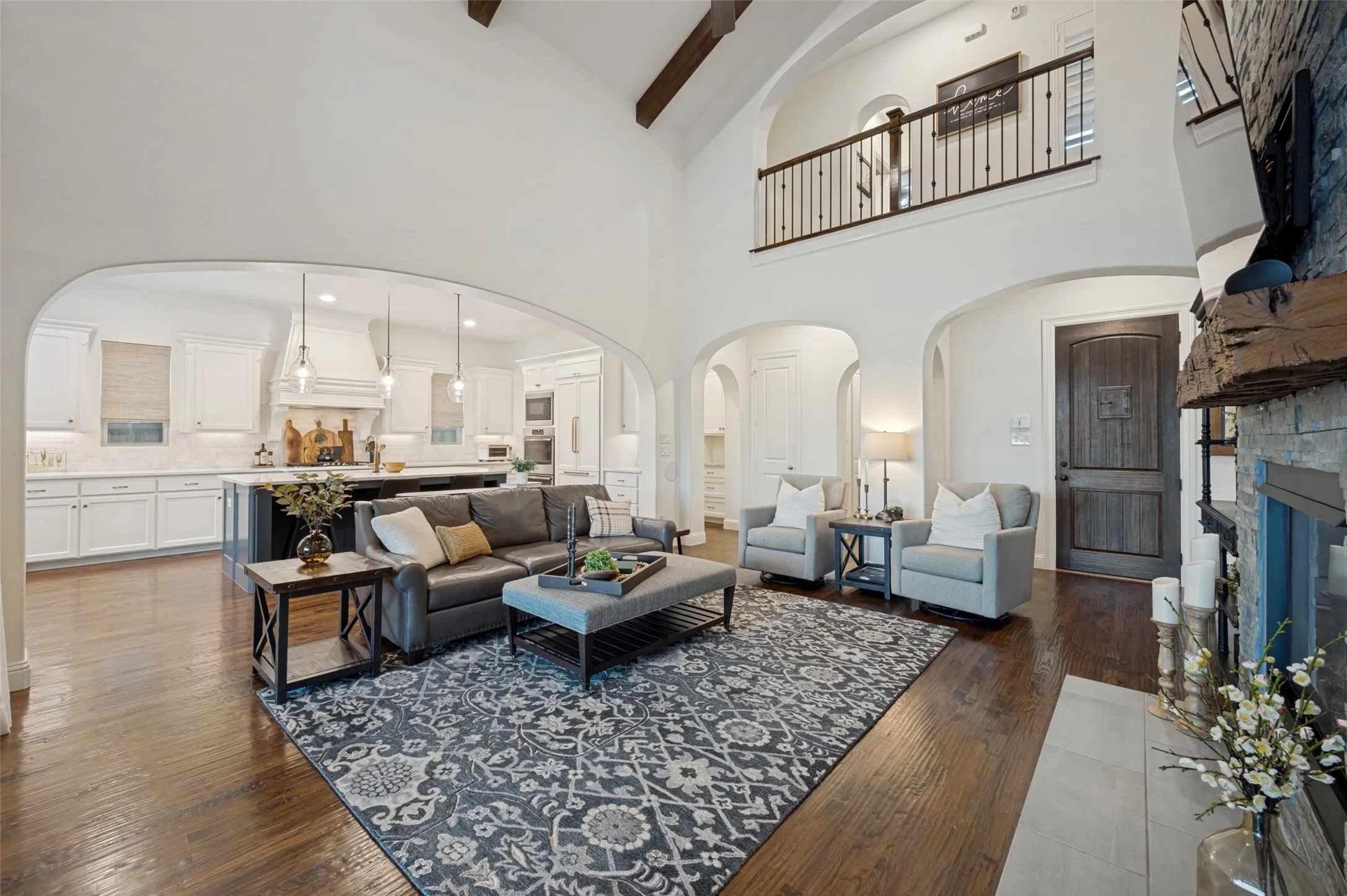 A grand living area features soaring vaulted ceilings, accented by rich wood beams, creating a beautiful sense of height and architectural character. Warm hardwood floors, graceful arched openings, built in Sonos sound, and a dramatic stone fireplace elevate the space.