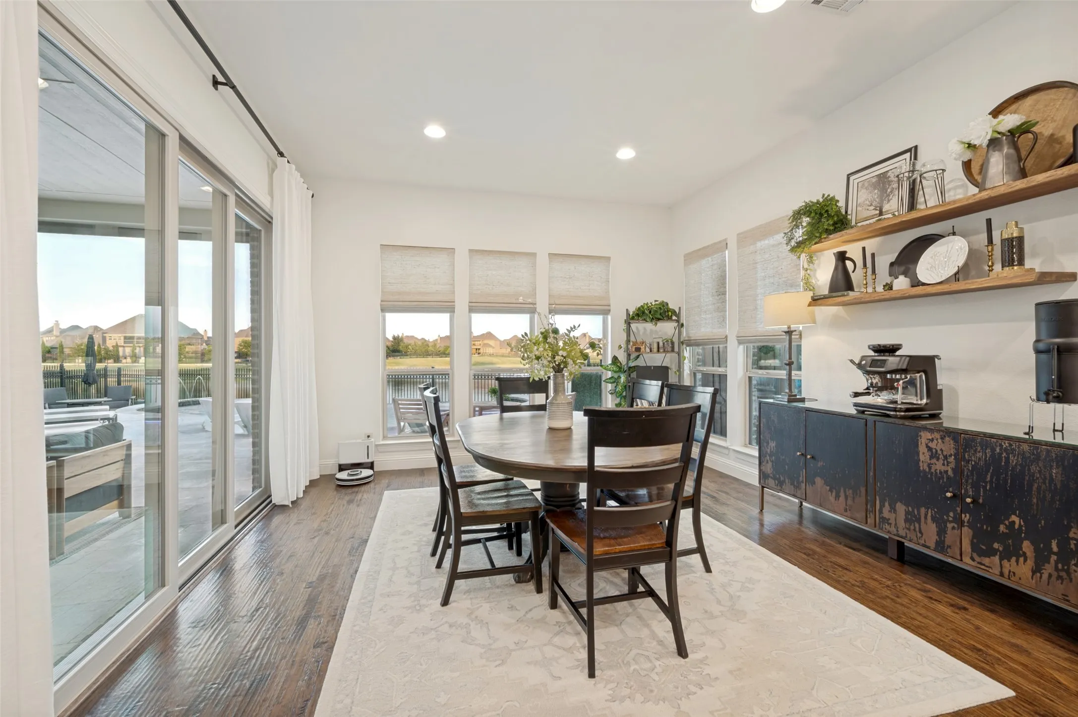 A bright and inviting dining area surrounded by windows with automatic shades, offering beautiful water and backyard views while providing effortless light and privacy control. Rich hardwood floors and custom walnut floating shelves add warmth and character.