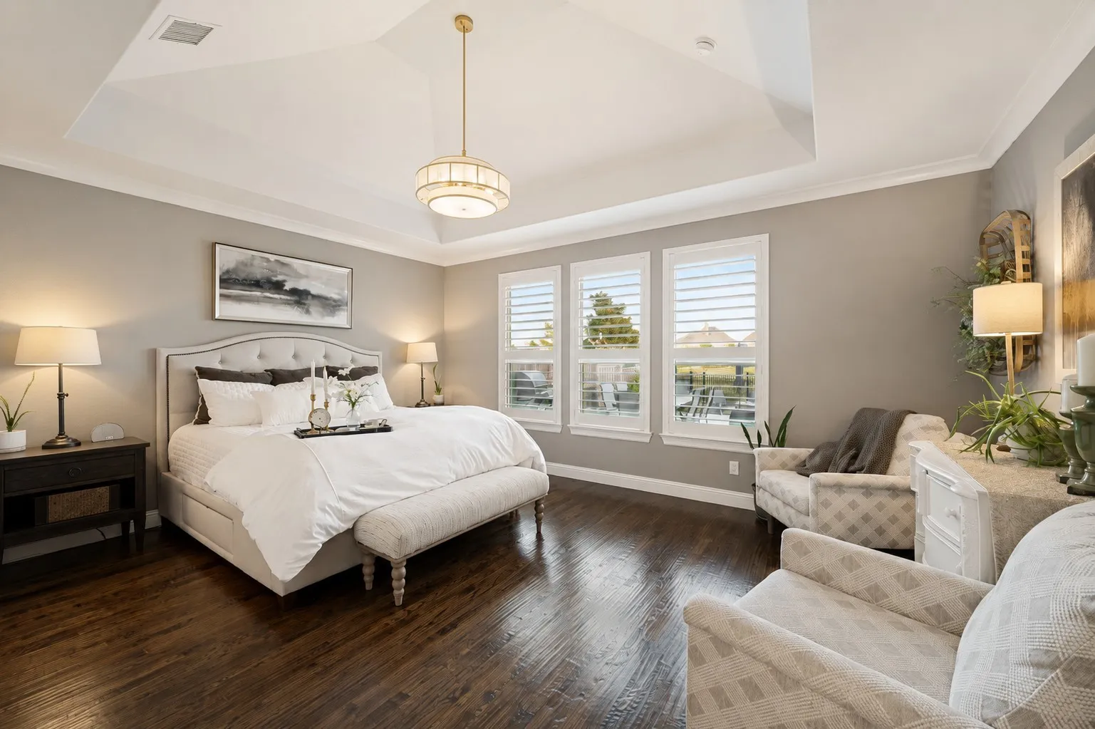 A serene and spacious primary suite offers the perfect private retreat, featuring rich hardwood floors, an elegant tray ceiling with designer lighting, and plenty of room for a comfortable sitting area. A wall of windows with plantation shutters fills the room with natural light while offering peaceful pond and fountain views, creating a relaxing setting to begin and end each day.