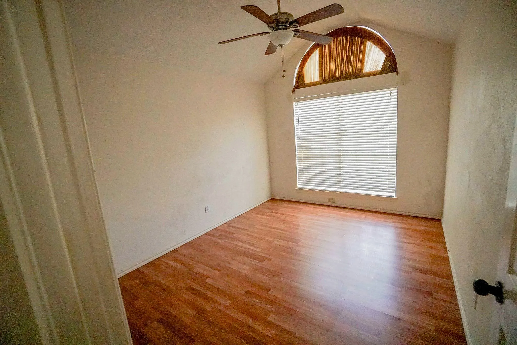 Vaulted ceiling, wood-finish flooring, ceiling fan, large window with blinds, and an arched transom window
