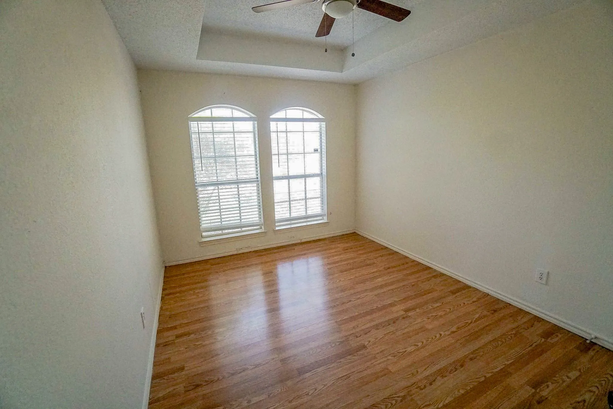 Wood-finish flooring, tray ceiling with ceiling fan, and two arched windows with blinds