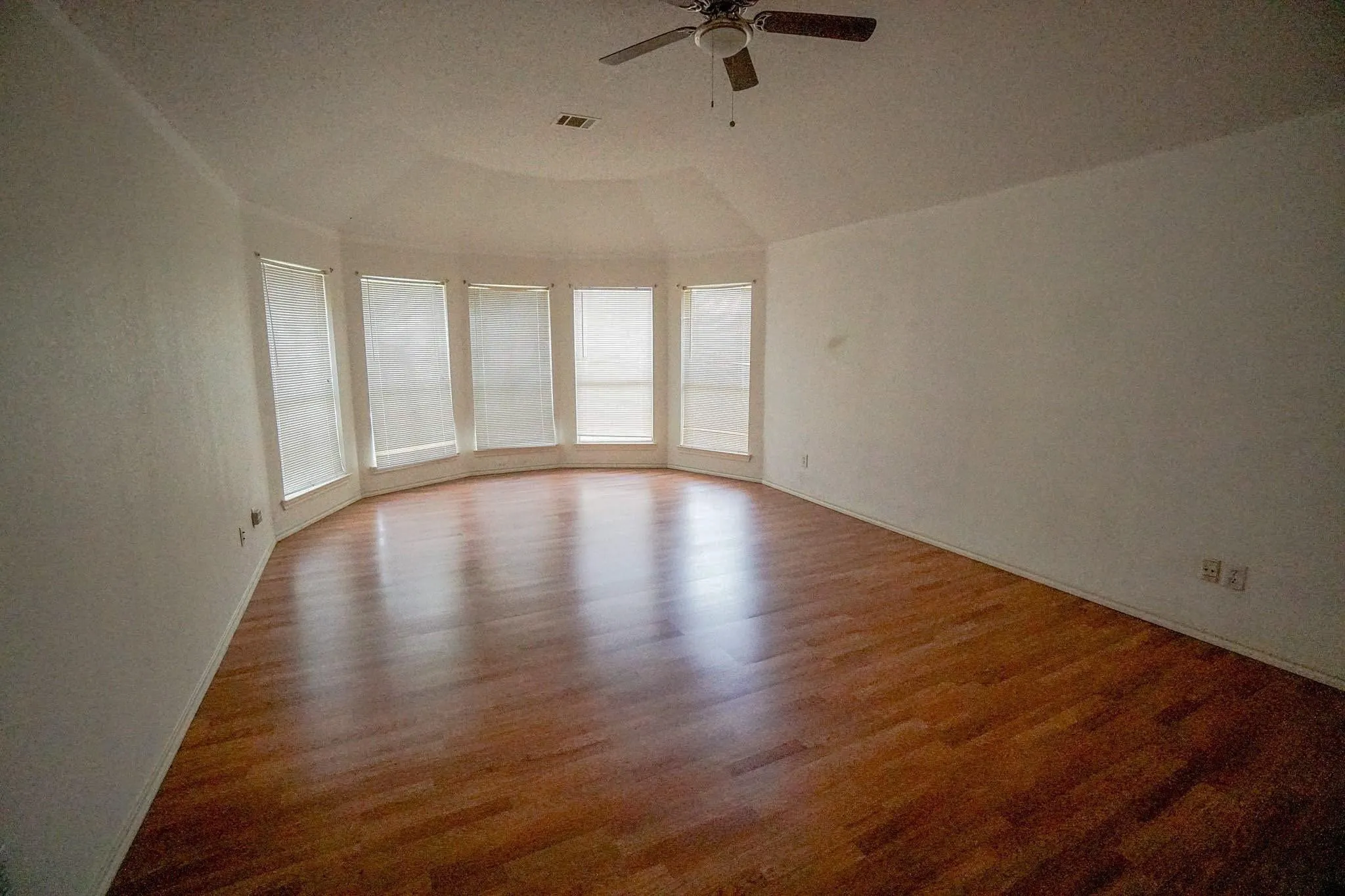 Spacious room with wood-finish flooring, a ceiling fan, and a curved bay window featuring multiple windows with blinds