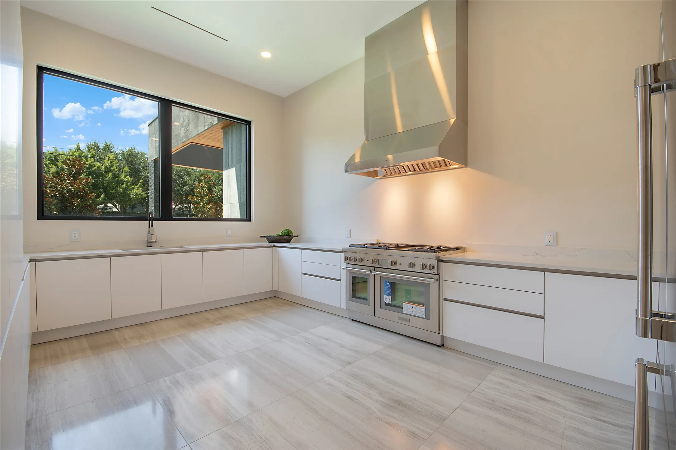 White flat-panel cabinetry with integrated pulls, stainless steel appliances, a professional-grade range with a substantial range hood, light-toned countertops, and large format floor tiles