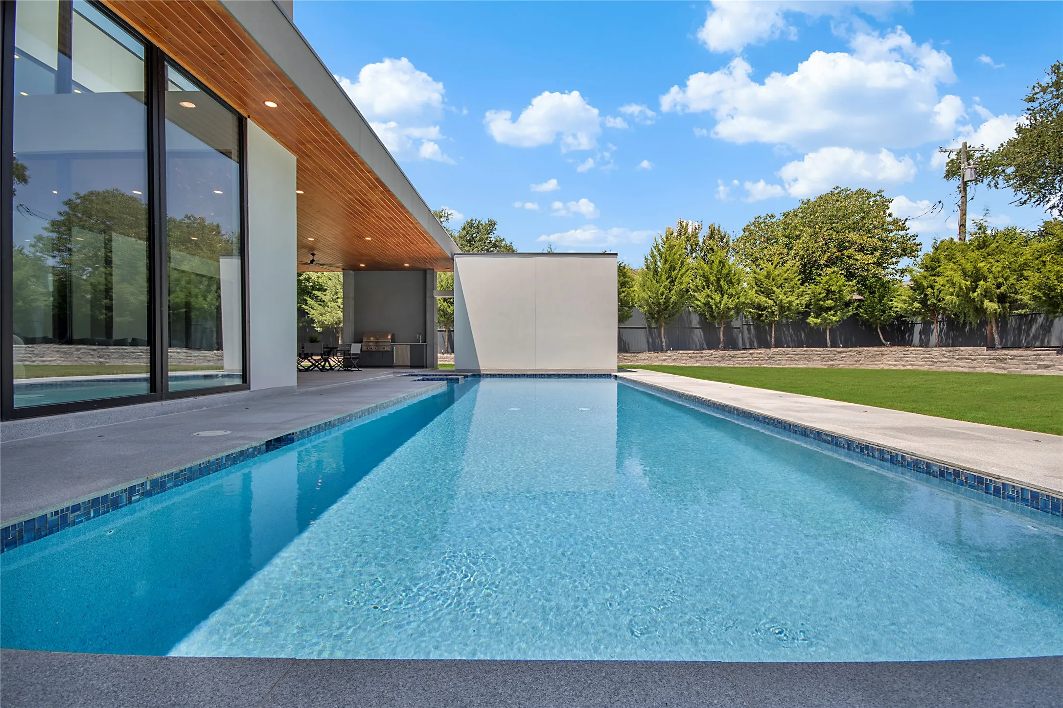 Expansive swimming pool with mosaic tile trim, a large grass yard, and an attached covered patio featuring an outdoor kitchen and wood-finish ceiling