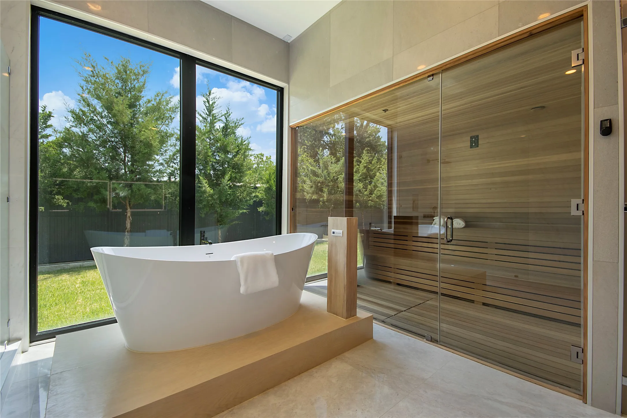 Freestanding soaking tub on a raised platform, floor-to-ceiling windows, natural wood sauna with glass enclosure, light-toned tile flooring, and a textured wall finish