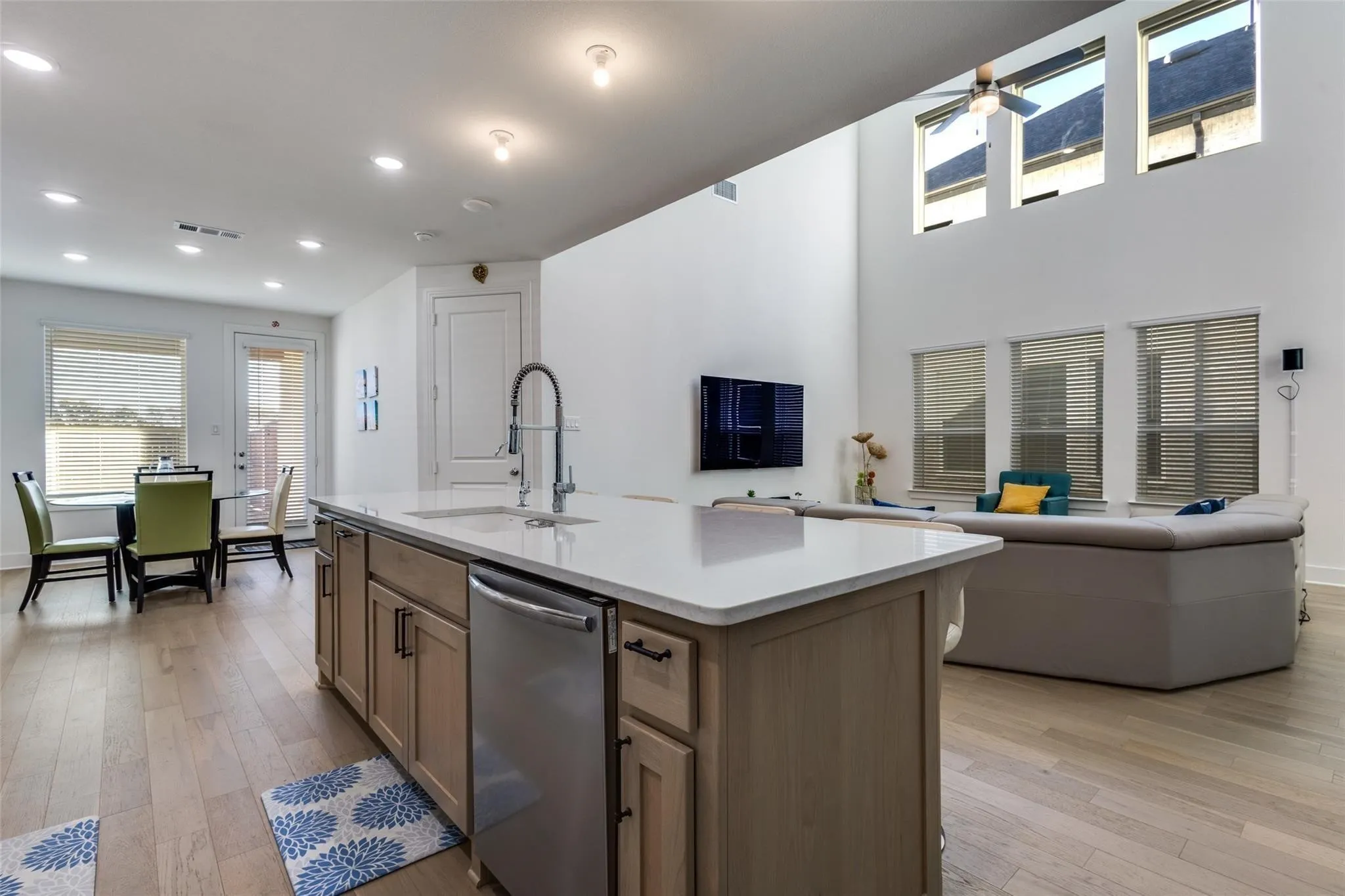 Open-concept living space with wood-finish flooring, a kitchen island with an integrated sink and dishwasher, high ceilings, and multiple windows with blinds
