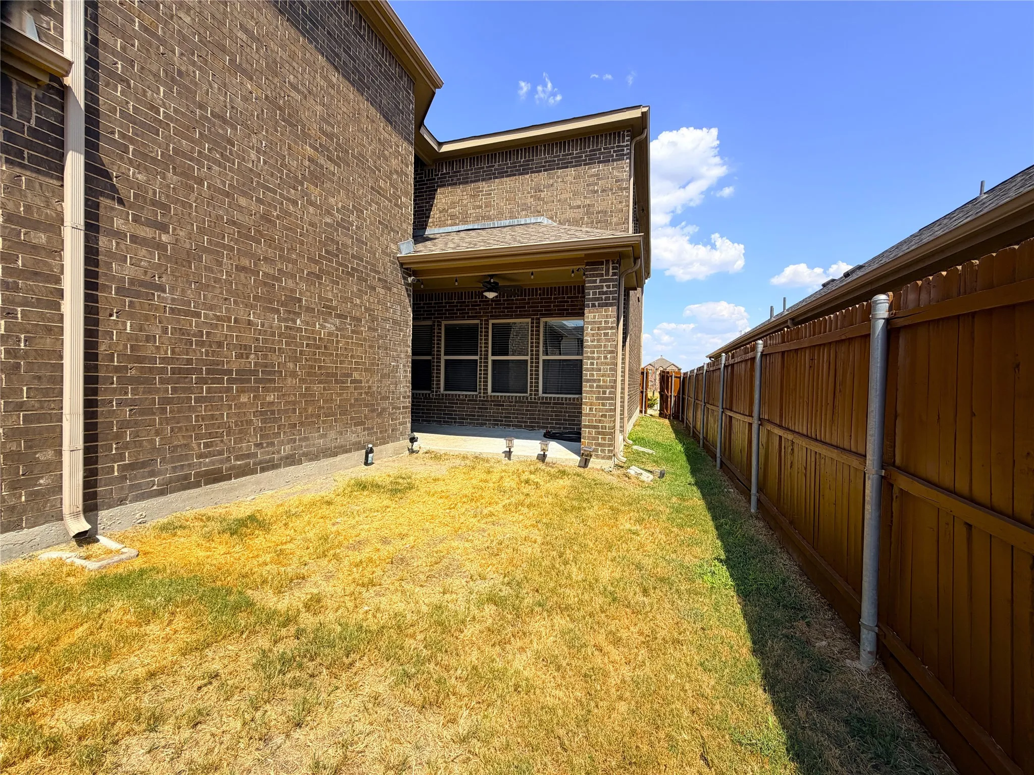 Spacious backyard with level lawn and wooden privacy fencing, adjoining a brick residence featuring a covered patio entry and oversized transom window beneath clear blue skies