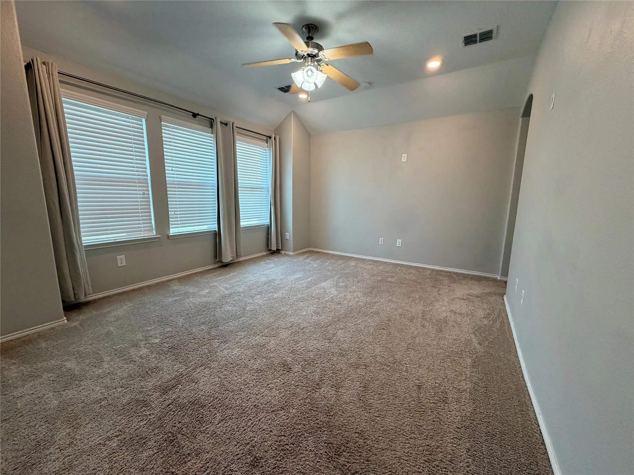 Spacious room featuring neutral carpet, multiple windows with blinds, a ceiling fan with integrated lighting, recessed lighting, and an arched doorway