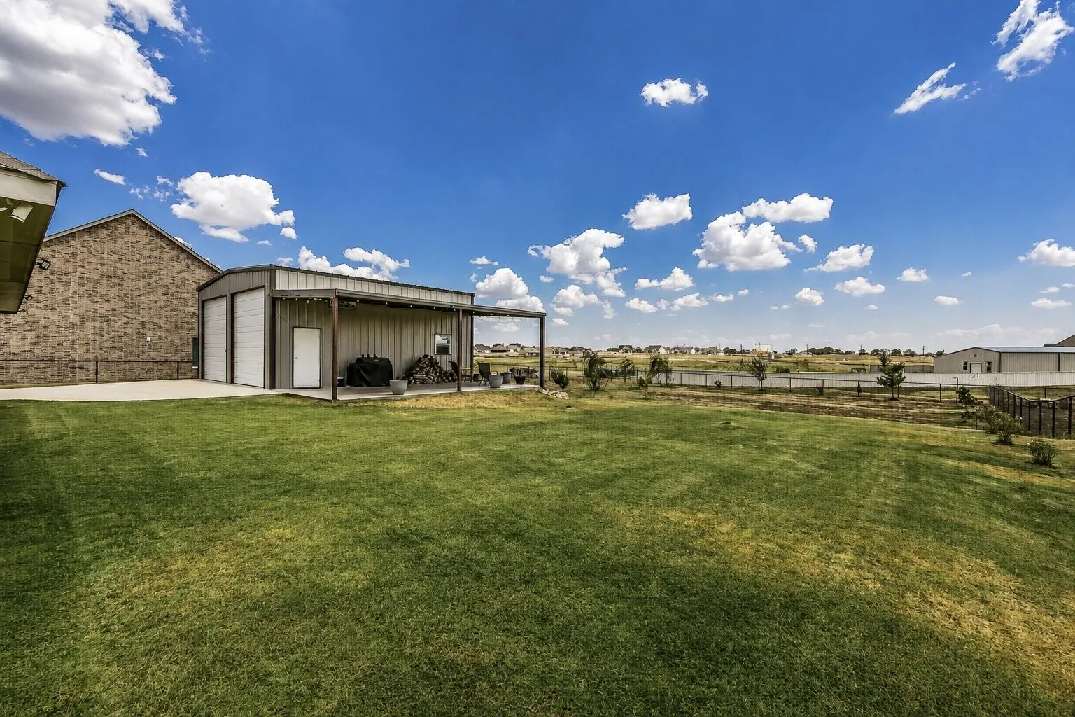 Expansive green lawn, brick exterior, detached metal building with two garage doors, covered outdoor area, and a white entry door