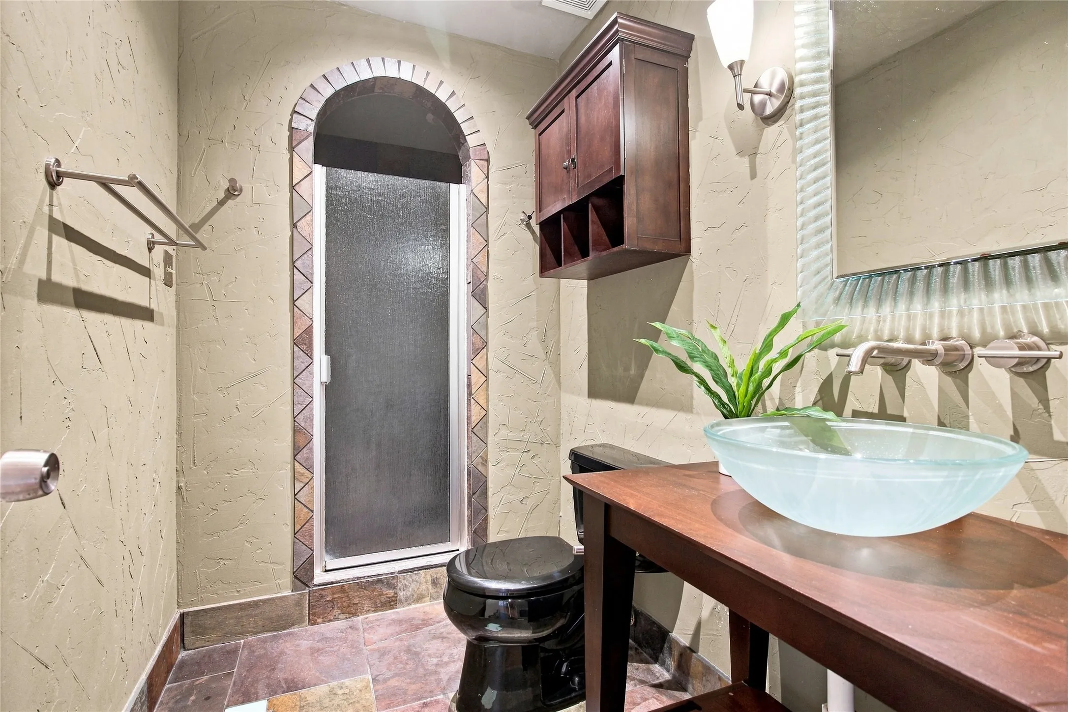 Bathroom with  wall finish, arched shower entry with decorative tile, glass vessel sink, wall-mounted faucet, and dark wood vanity with cabinet storage