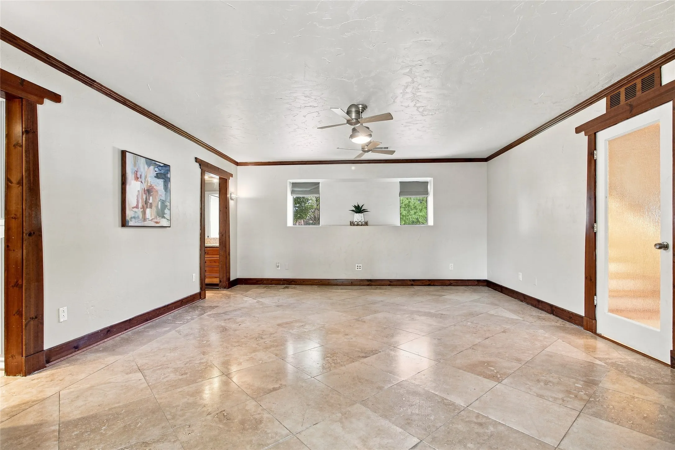 Another view of the primary bedroom with neutral-toned tile flooring, two ceiling fans, white walls with dark wood trim, and a door with a frosted glass panel