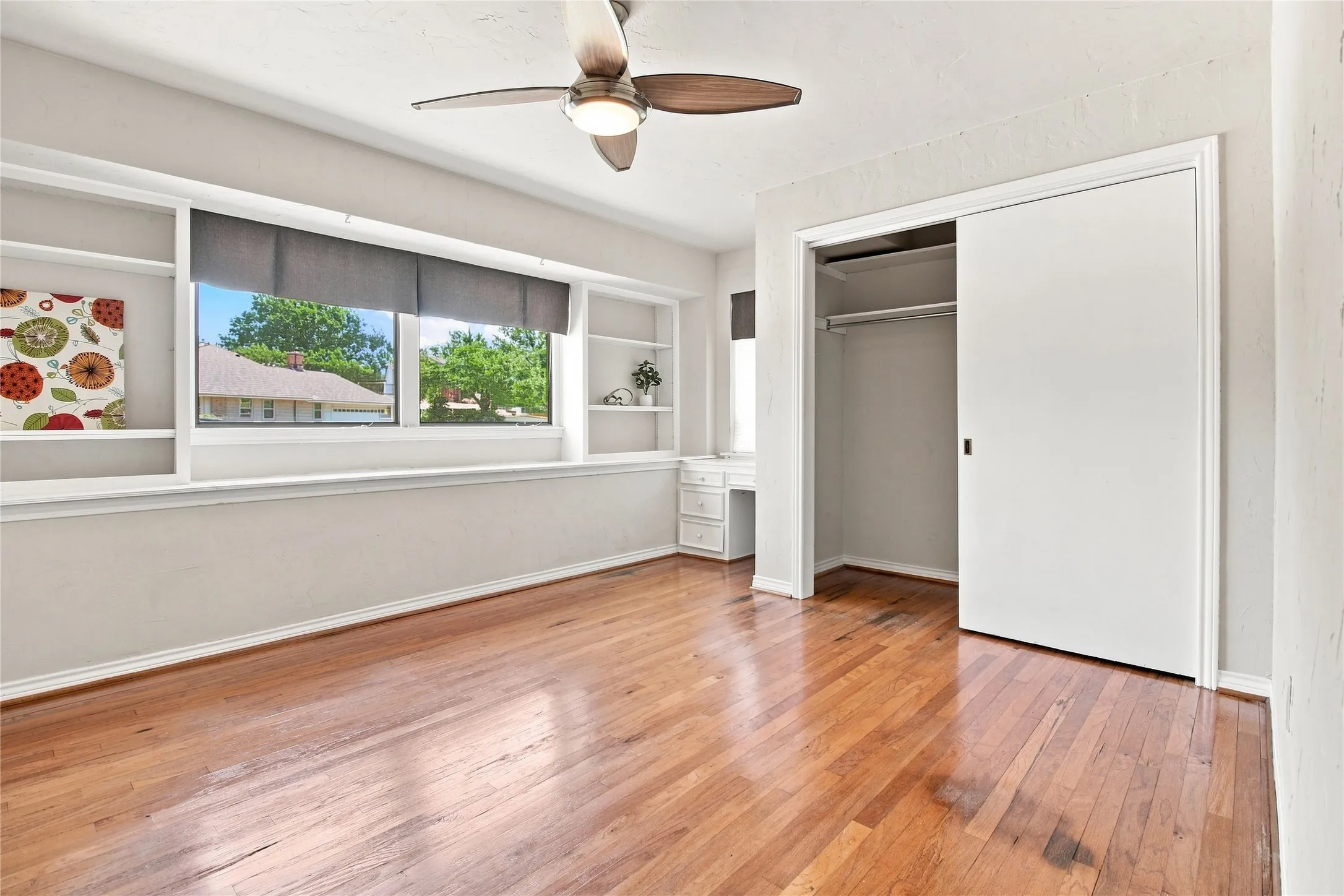Upstairs guest bedroom with hardwood flooring, built-in shelving, window with roller shades, built-in desk, and ceiling fan
