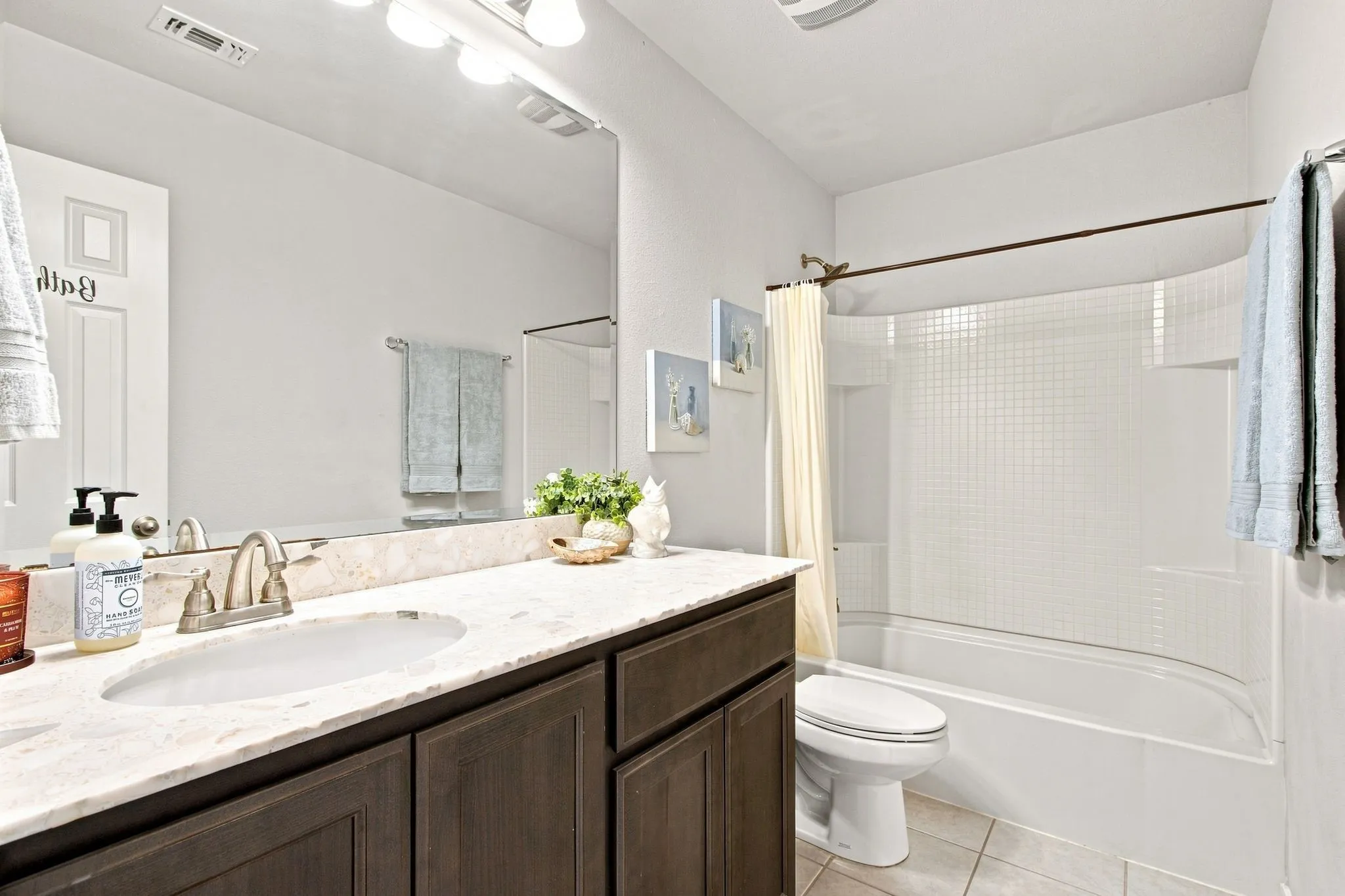 Bathroom featuring a wood-finish vanity with a light stone countertop, an integrated sink, a large wall-mounted mirror, and a white shower-over-tub combination.