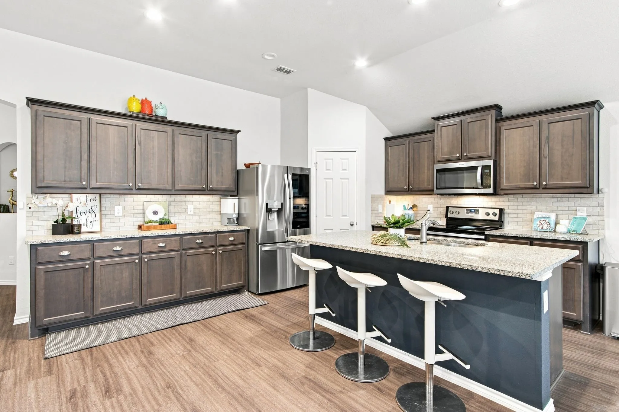 Spacious kitchen featuring an expansive island, wood-finish LVP flooring, stainless steel appliances, dark wood cabinetry, and a subway tile backsplash