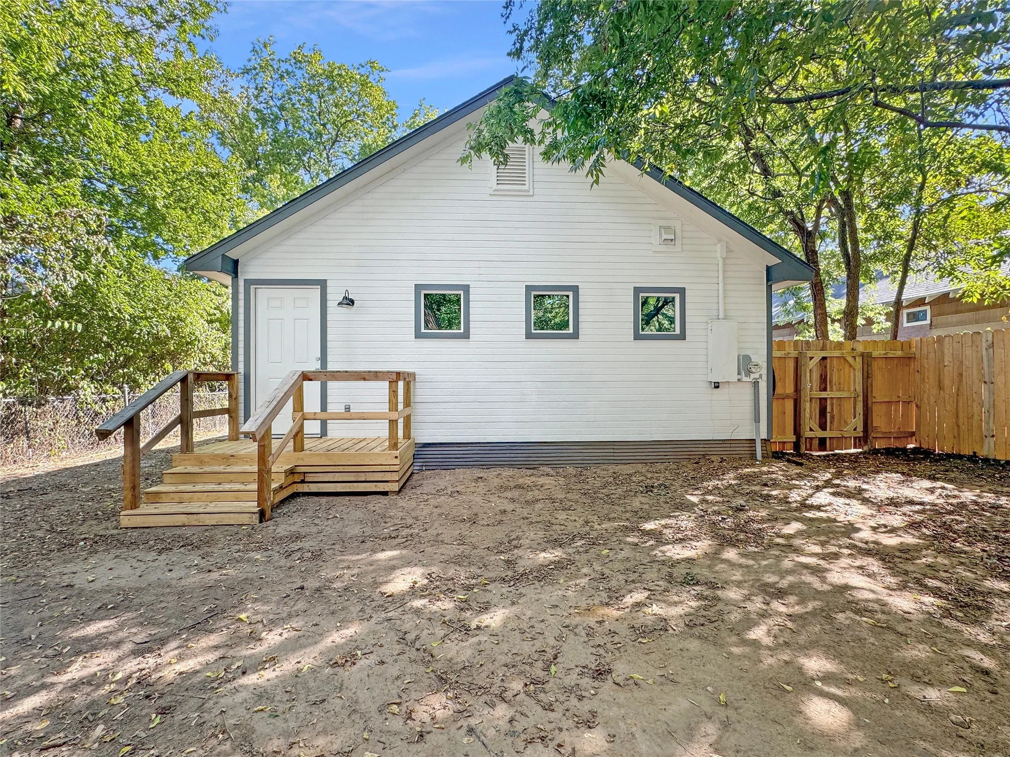 White siding exterior with a dark gray trim accent, featuring a wooden deck and steps, a wooden fence, and multiple small windows