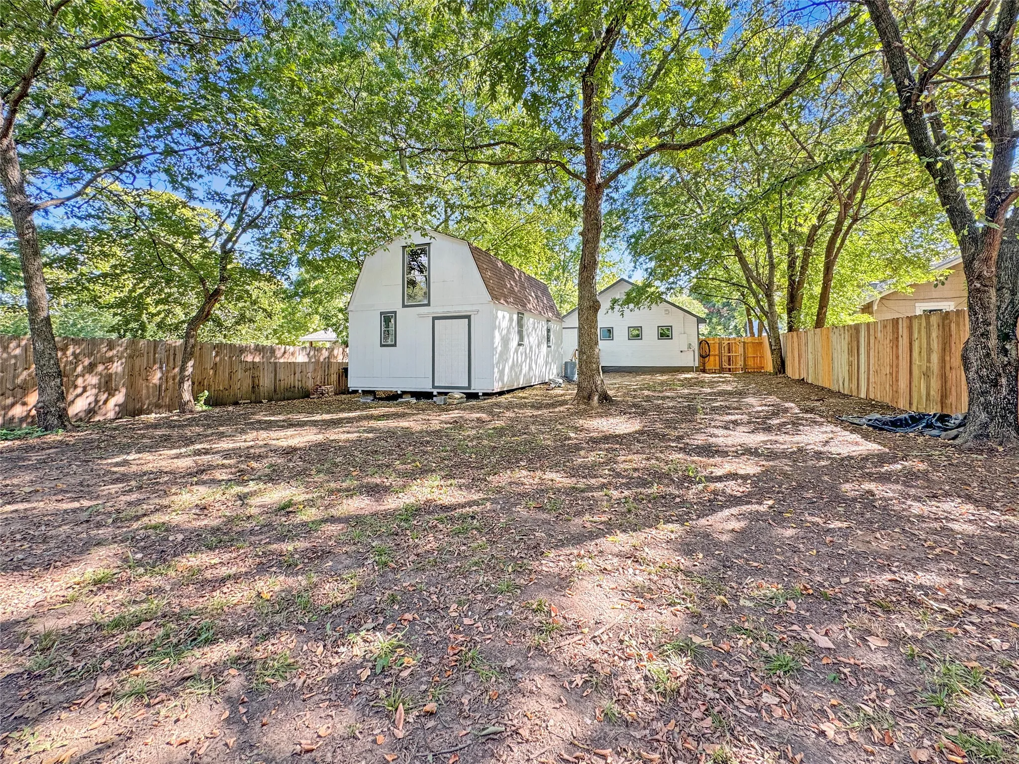 Spacious backyard featuring a white storage building/barn with a gambrel roof and multiple windows, mature trees, and privacy fencing