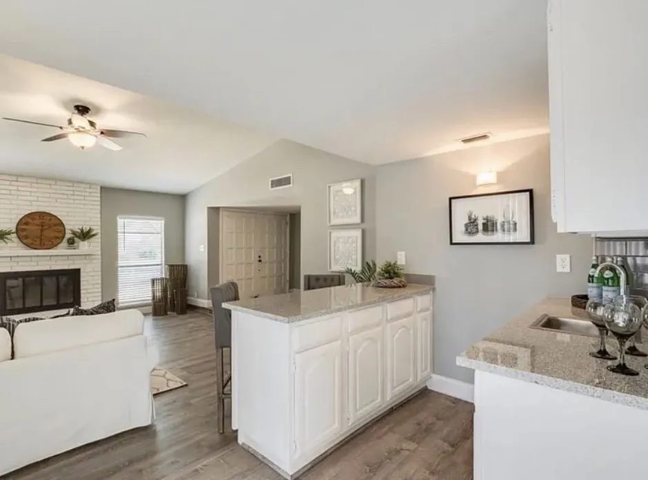 Open-concept kitchen and living area featuring white cabinetry with raised-panel doors, granite countertops, a large center island with seating, stainless steel appliances, wood-finish flooring, and vaulted ceilings with recessed lighting