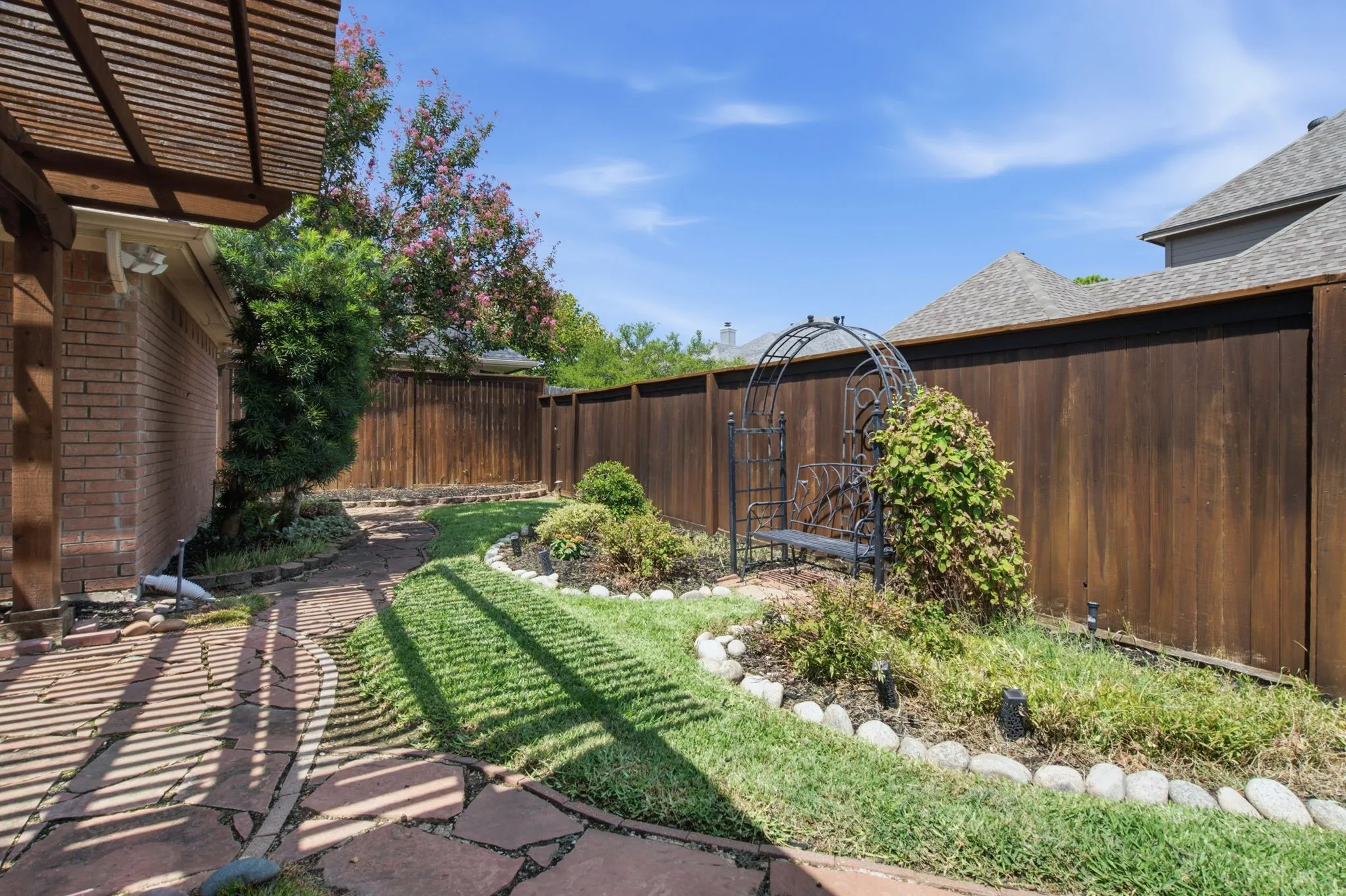 Expansive backyard oasis featuring a manicured lawn, mature landscaping with shrubs and flowering plants, wood-finish privacy fencing, and a metal arbor structure