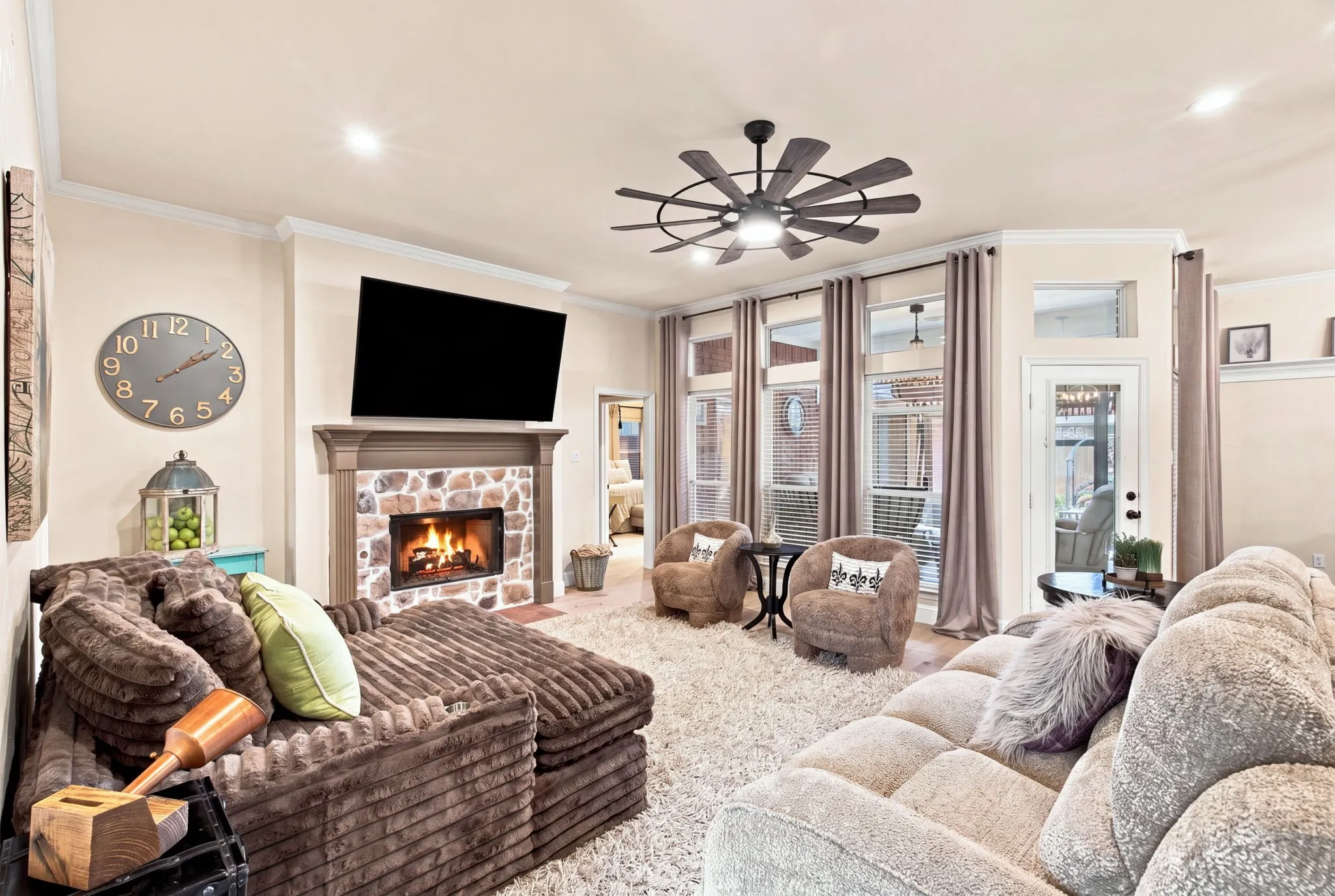 Stone-clad fireplace with mantel, crown molding, a multi-blade ceiling fan, and multiple windows with transoms