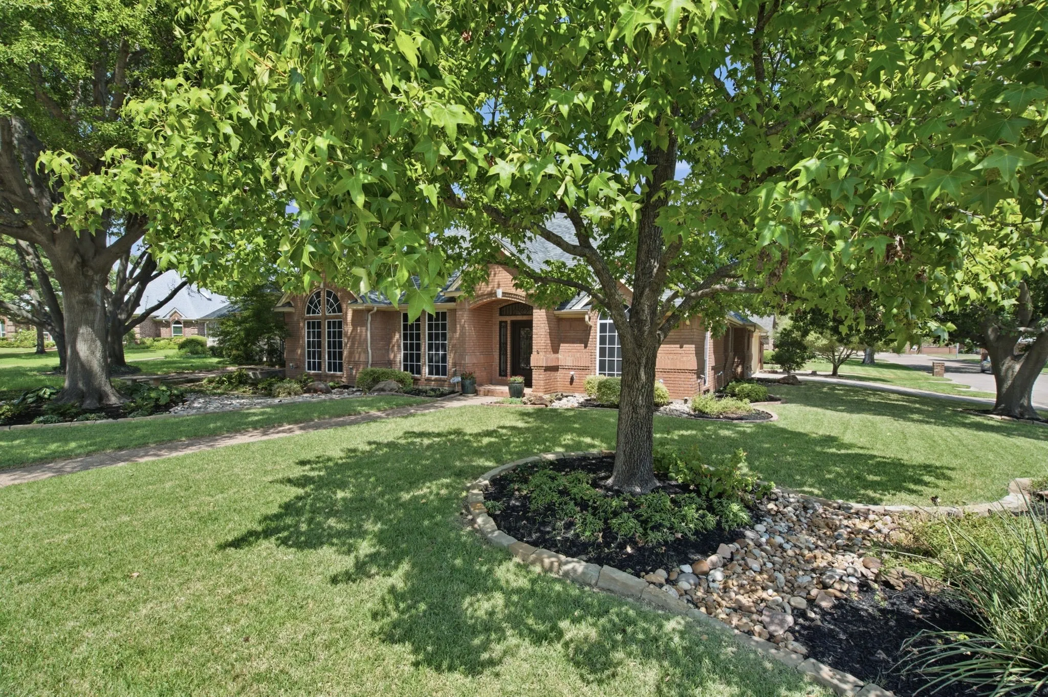 Brick exterior facade with arched window details, mature trees, manicured lawn, and a decorative dry creek bed with stone edging