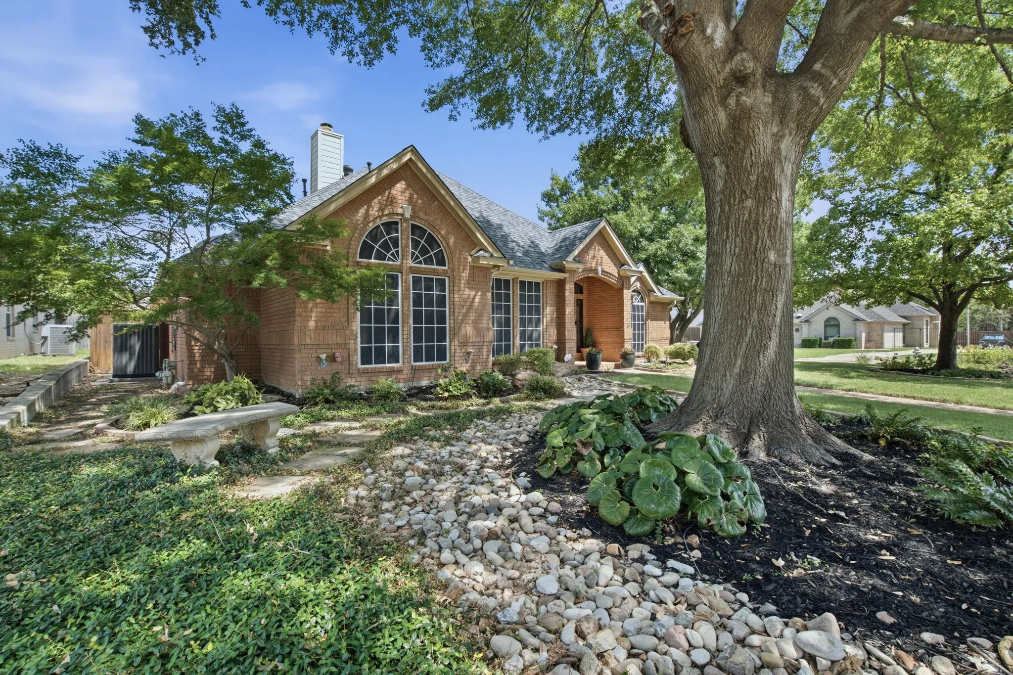 Brick exterior with arched windows, prominent chimney, mature trees, and stone hardscaping