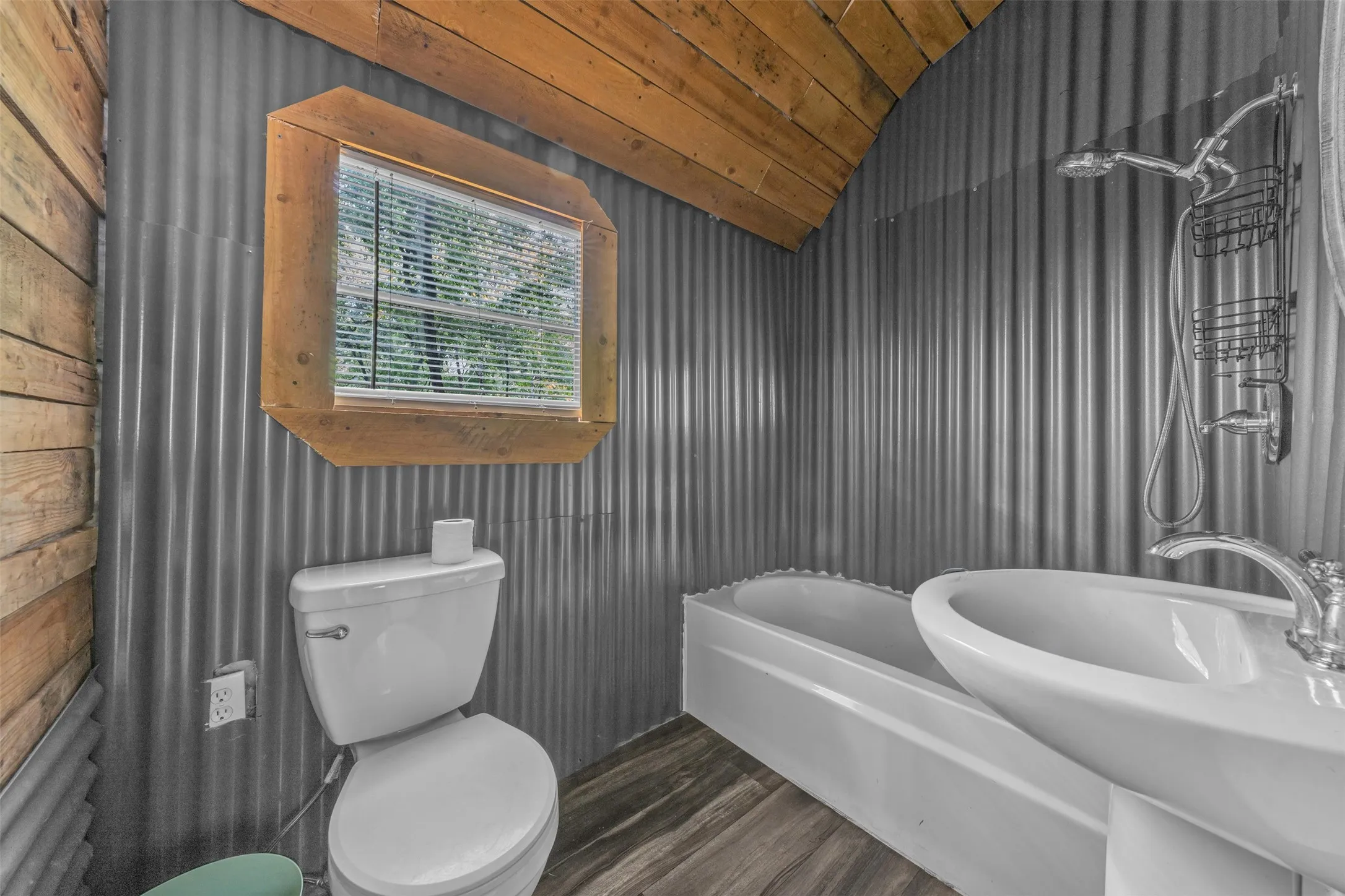 Corrugated metal walls featuring a wooden-framed window, a white porcelain toilet, a white drop-in bathtub, and wood-finish flooring