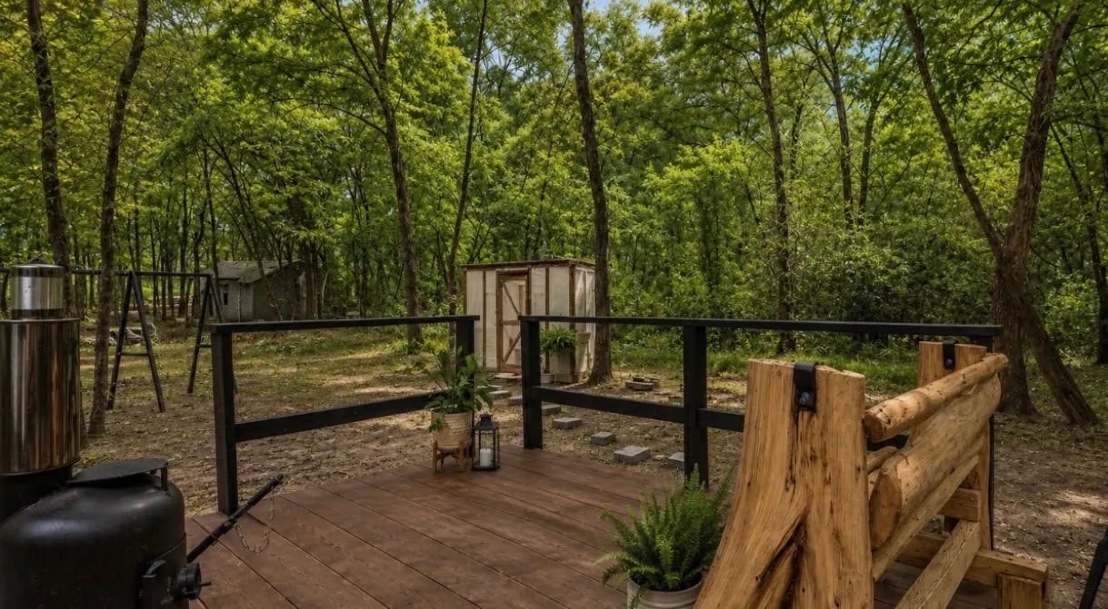 Wood-finish deck with dark railing, surrounded by mature trees, featuring a rustic wooden bench
