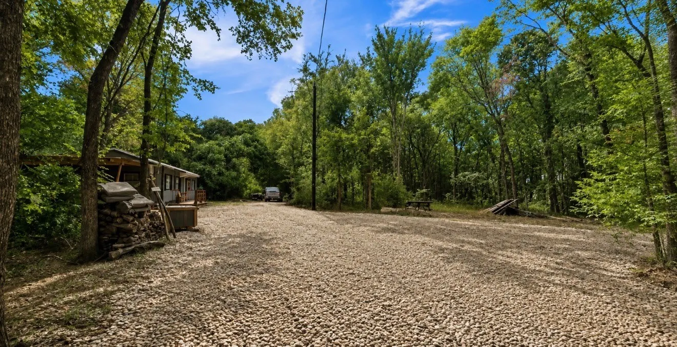 Wooded property with a gravel driveway, mature trees, and a visible structure with a porch