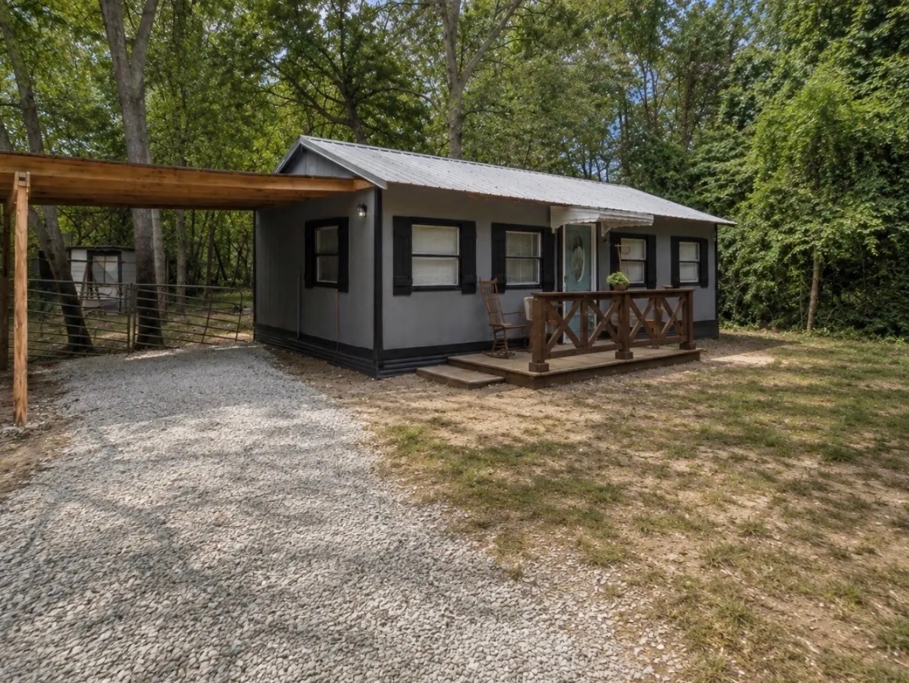 Gravel driveway leading to a covered carport, offering a gray exterior with black trim, a covered porch with a wood railing, and a metal roof