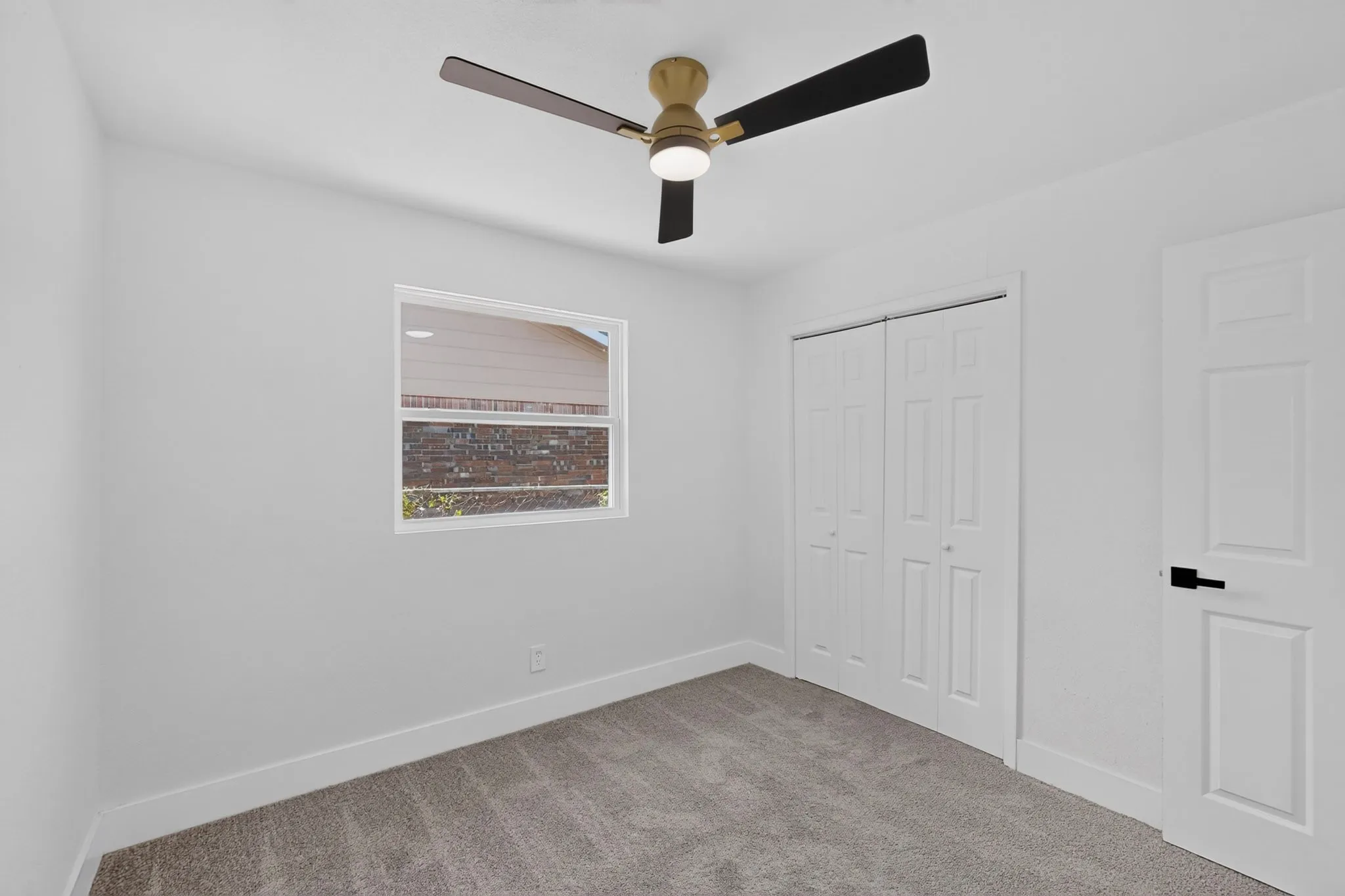 Neutral wall tones, gray carpeting, a window with a white frame, white bi-fold closet doors, and a ceiling fan with black blades