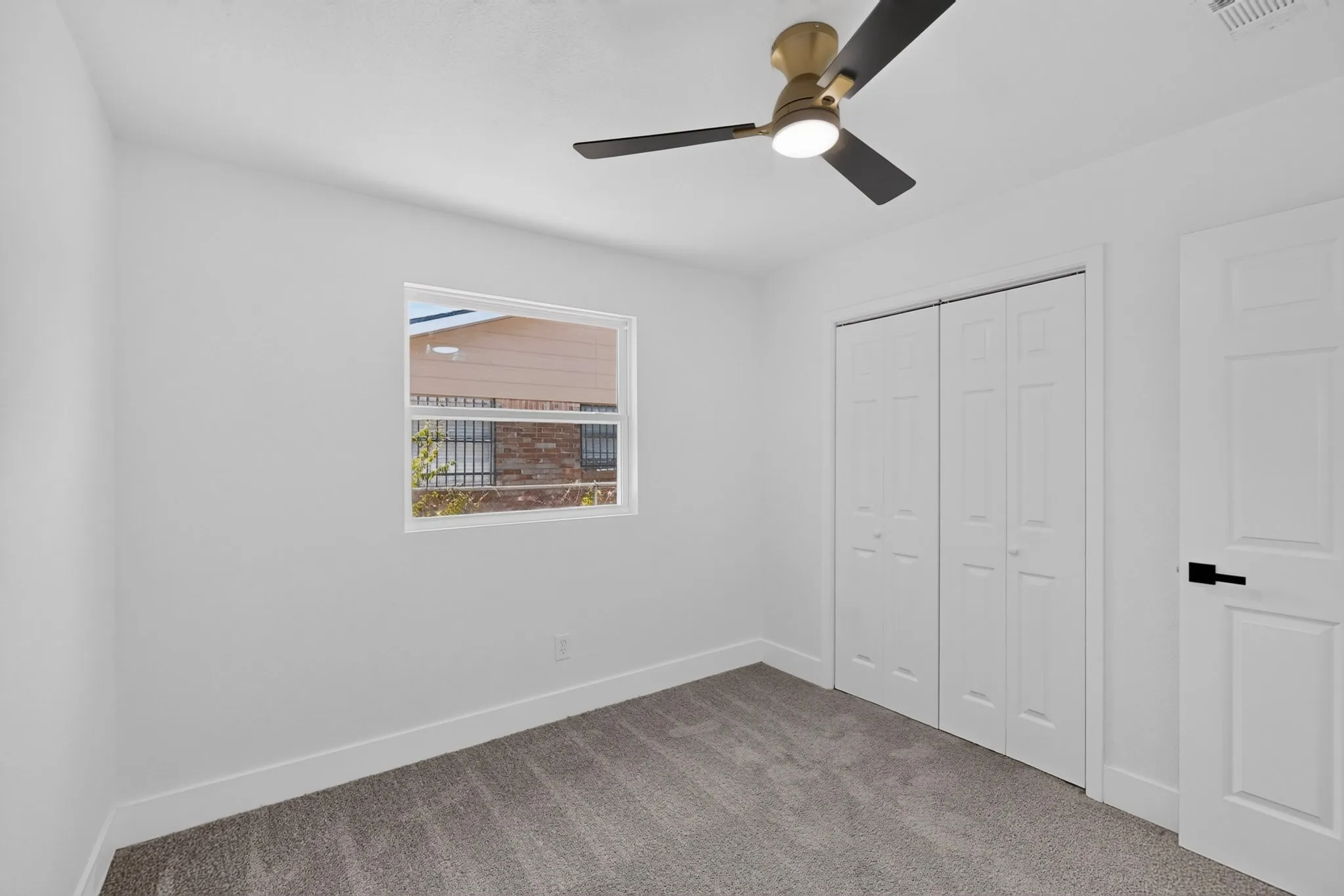 Crisp white walls, gray carpet flooring, a window with metal grates, a ceiling fan with light, and white paneled doors