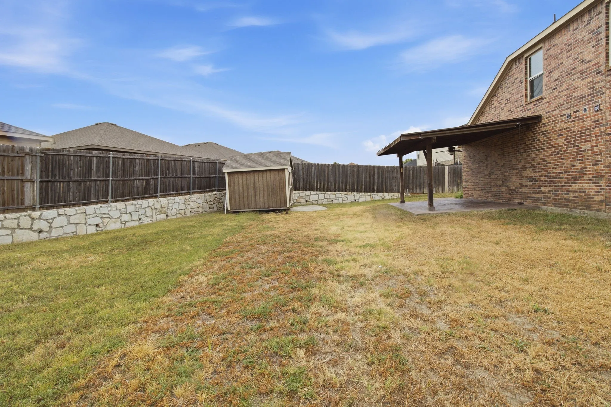 Spacious backyard featuring a covered patio, brick exterior, wood privacy fencing, a stone retaining wall, and a storage shed